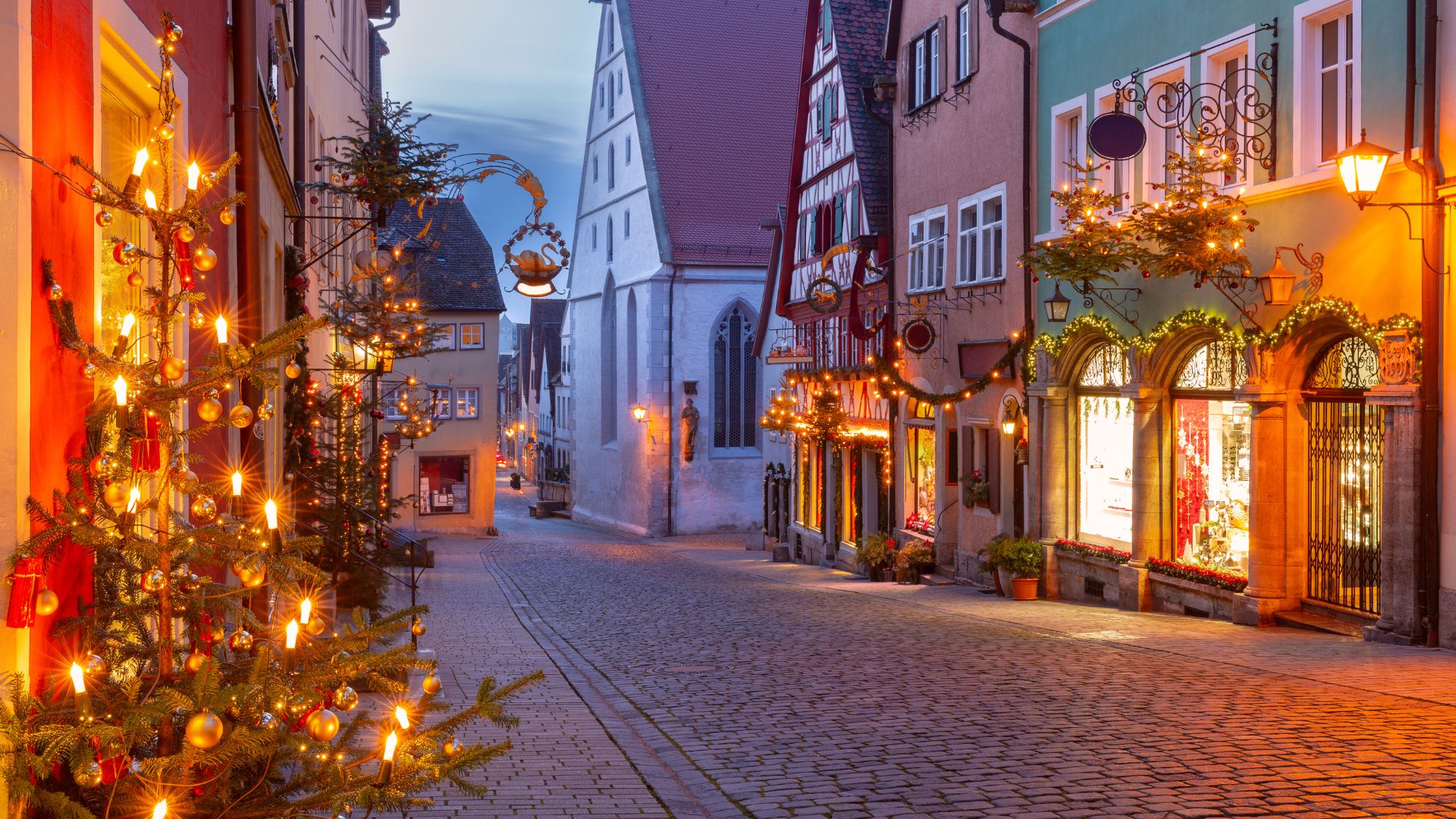 A cobblestone street in a European town is decorated with Christmas lights and trees at dusk.