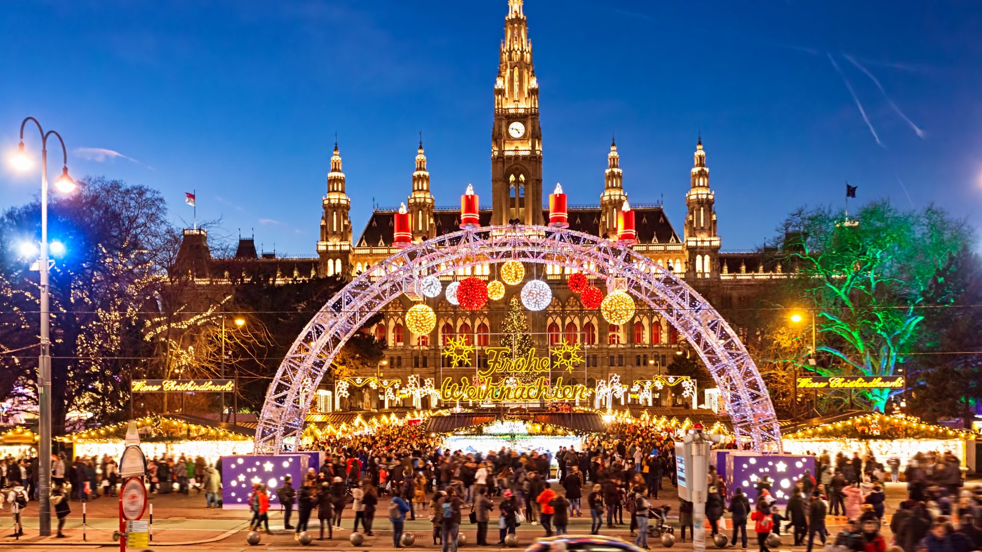 A crowded, brightly lit Christmas market in Vienna's Rathausplatz in front of the illuminated city hall building at dusk.
