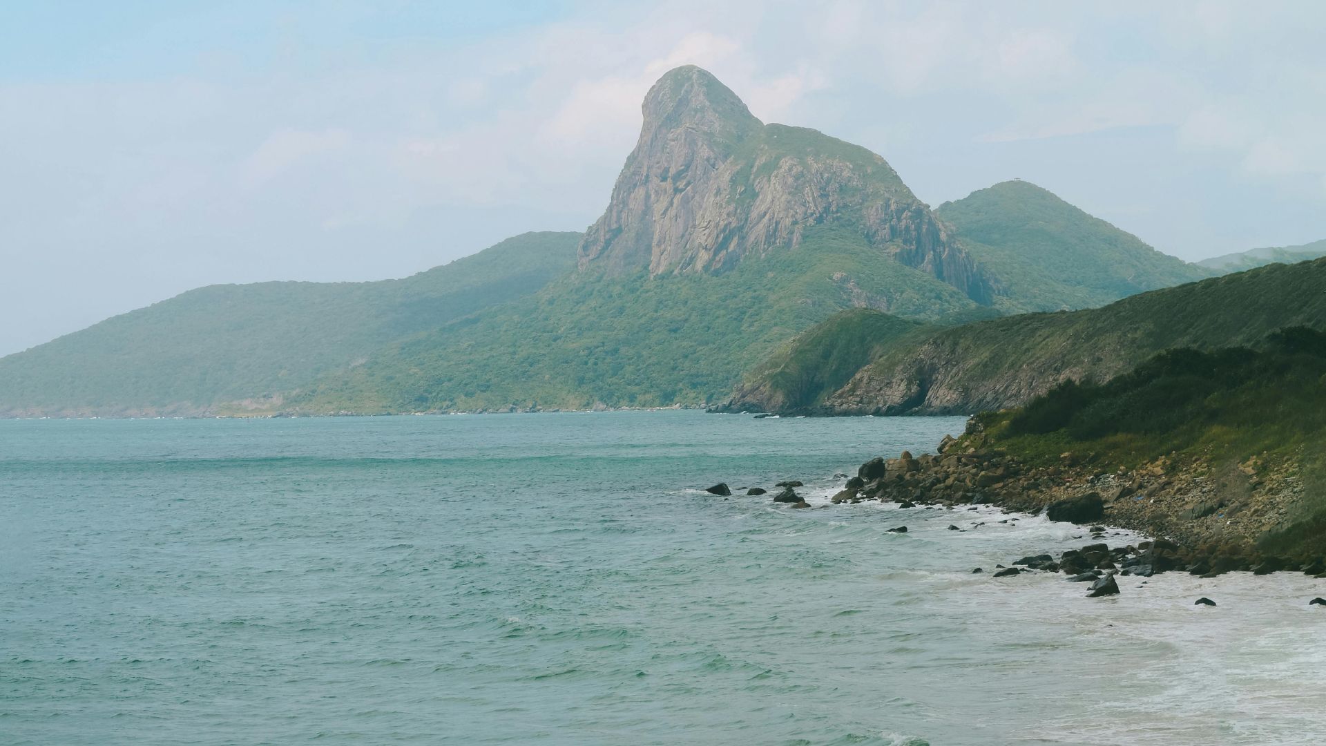 A beautiful coastal landscape photograph of the Con Dao Islands in southern Vietnam, featuring a rocky shoreline, turquoise water, and a prominent, lush green, jagged mountain peak rising in the background under a blue sky with white clouds.