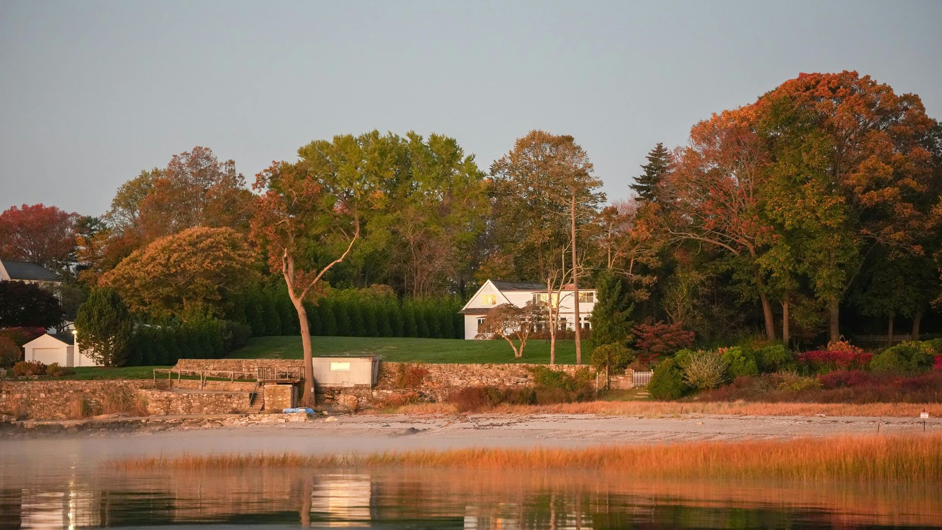 A large, elegant white house with a stone retaining wall sits on a green lawn above a sandy beach and a body of water. The surrounding trees show vibrant autumn foliage in green, orange, and red under a pale blue sky.