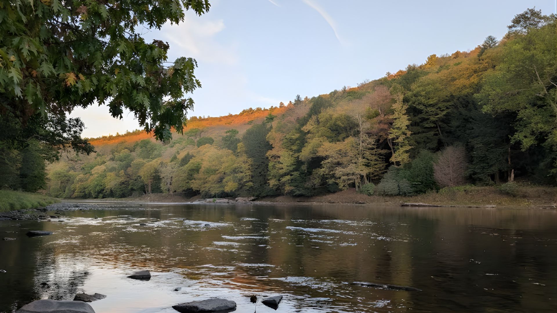 A river in the foreground with exposed rocks flows past a steep bank of forested hills displaying early autumn colors under a pale blue sky.