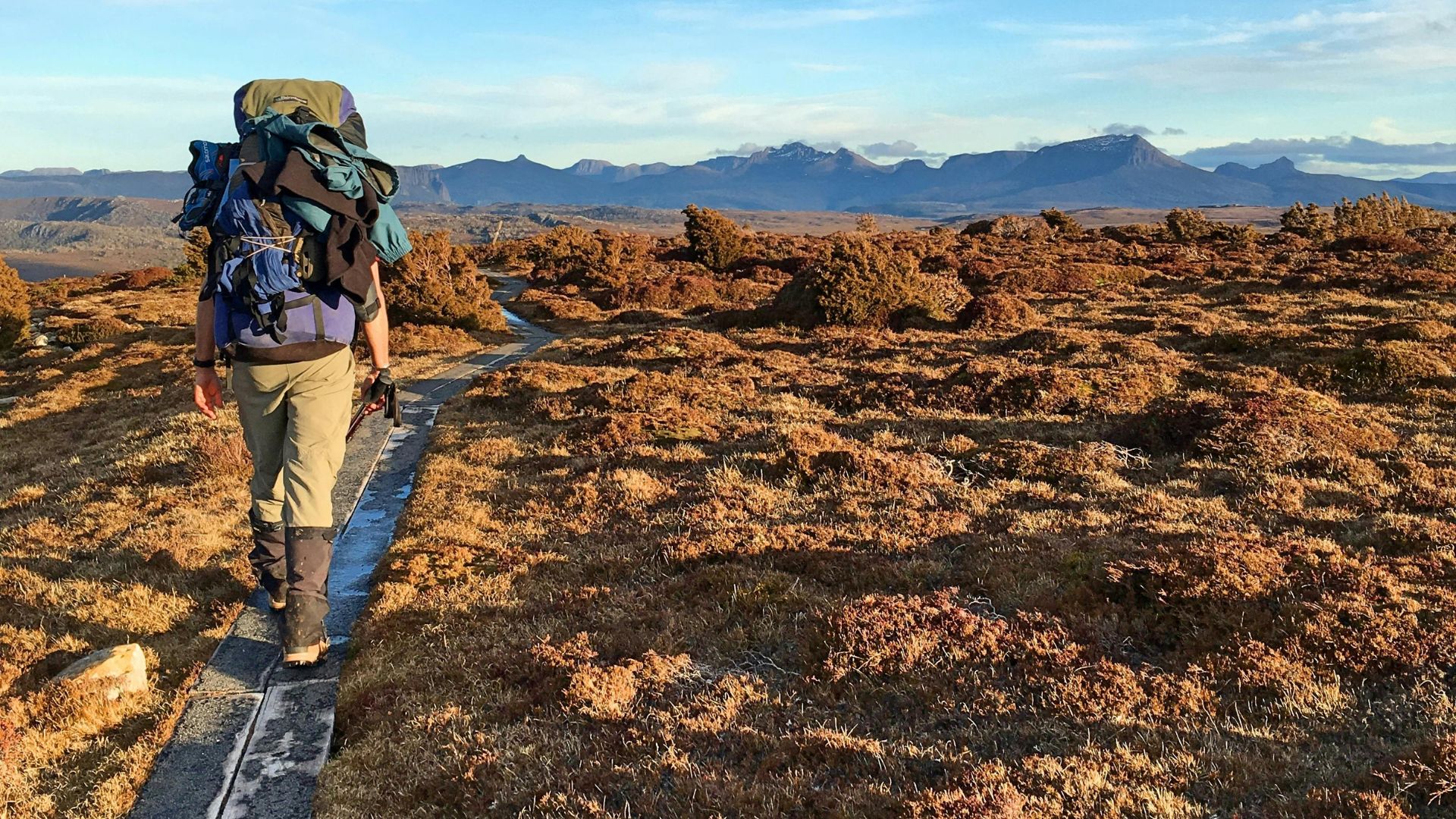 A hiker with a large backpack walks along a wooden boardwalk through a rugged, grassy alpine landscape toward distant mountains under a blue sky.