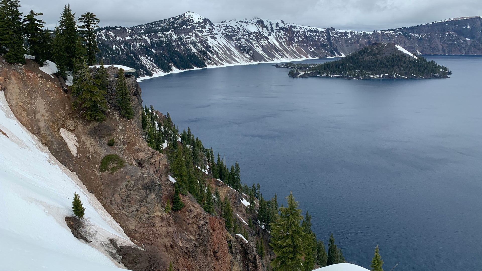 A view of a large, deep blue crater lake with a forested island and snow-covered cliffs under a cloudy sky.