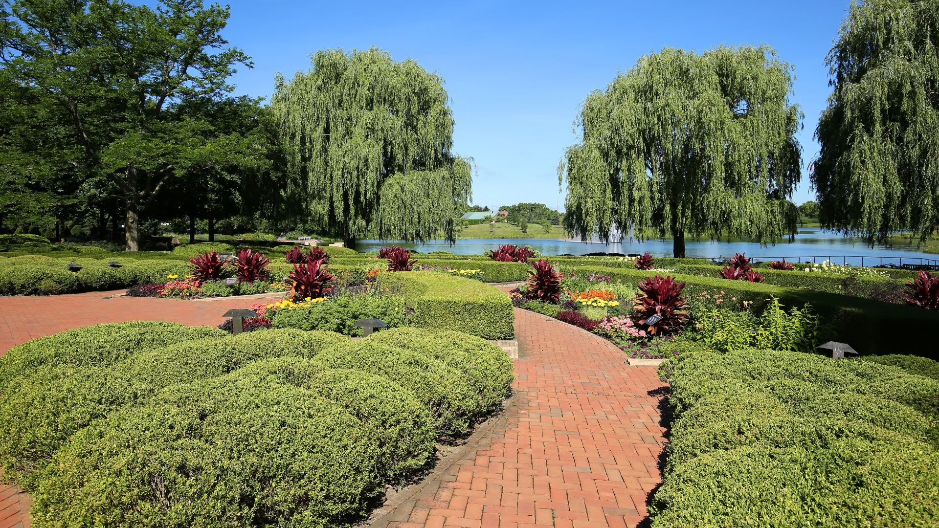 A brick pathway curves through formal, terraced garden beds featuring manicured green hedges and red and yellow flowers, leading toward a lake bordered by large weeping willow trees under a clear blue sky.