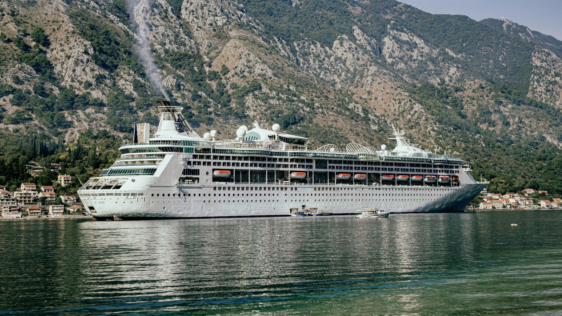 A large traditional mega-ship cruise ship is docked in a scenic bay surrounded by steep, green mountains.