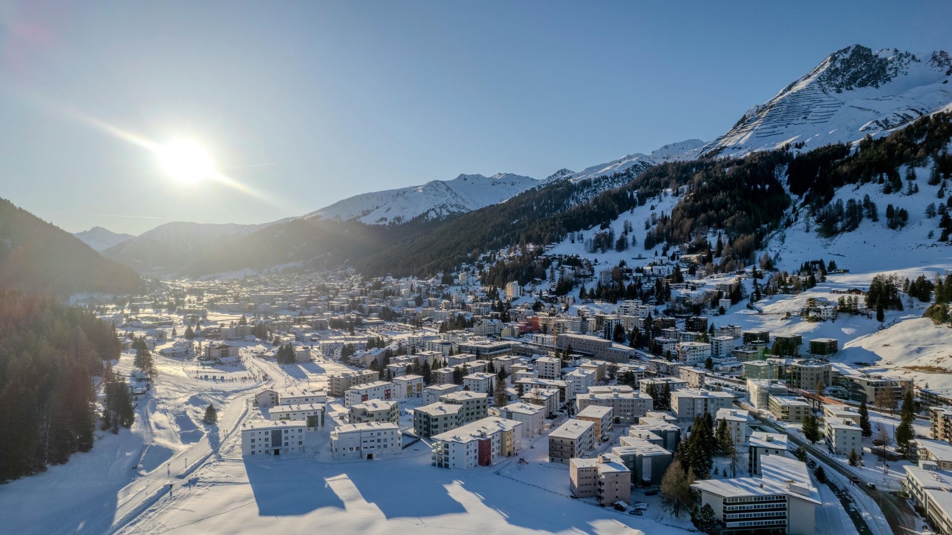 A picturesque mountain town nestled below snow-covered peaks under a clear blue sky. The town is surrounded by dense forests and expanses of snow.