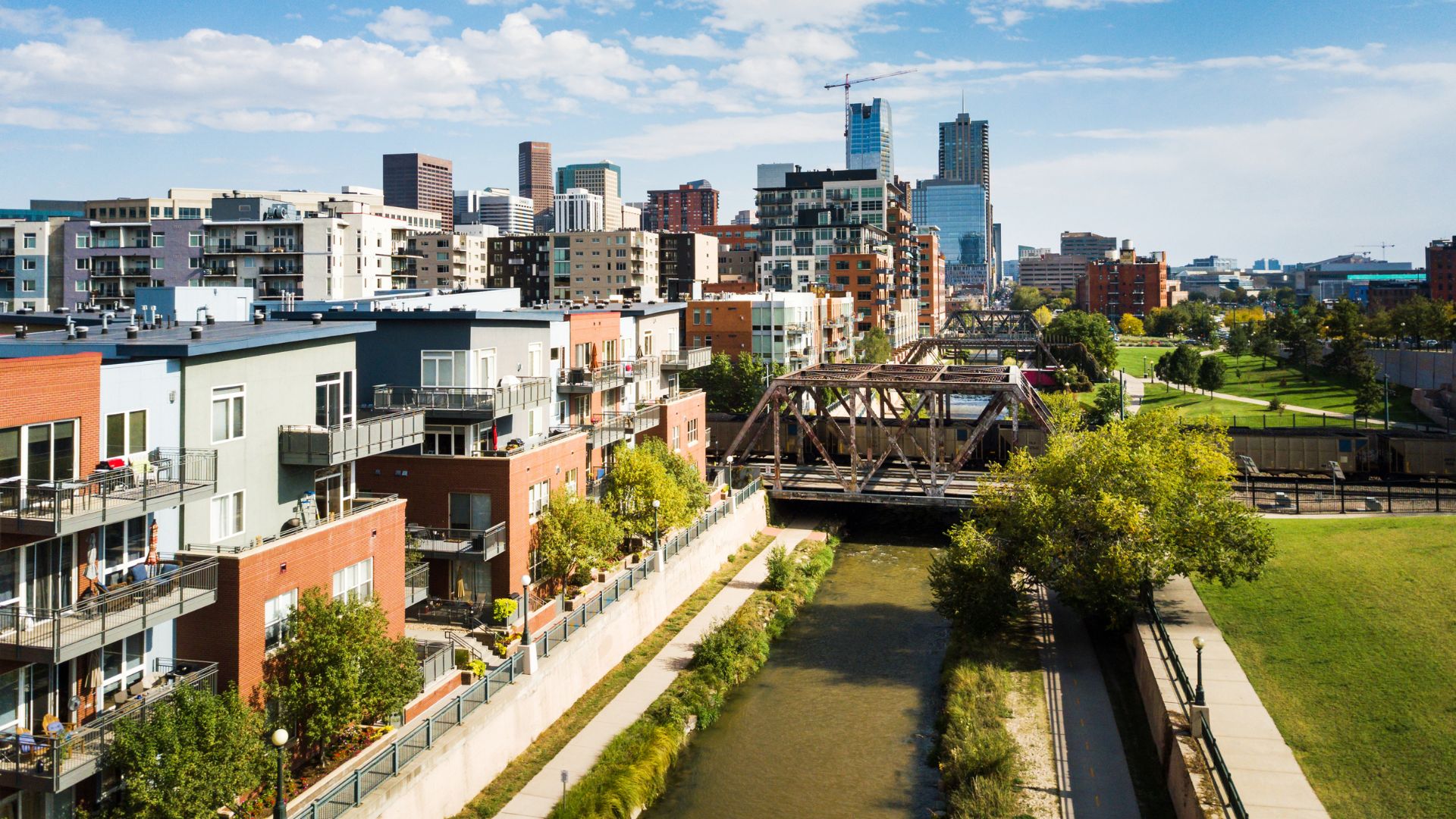 A panoramic daytime view of the Denver, Colorado city skyline featuring modern residential buildings next to a small river and a historic metal truss bridge, with downtown skyscrapers in the background under a partly cloudy sky.