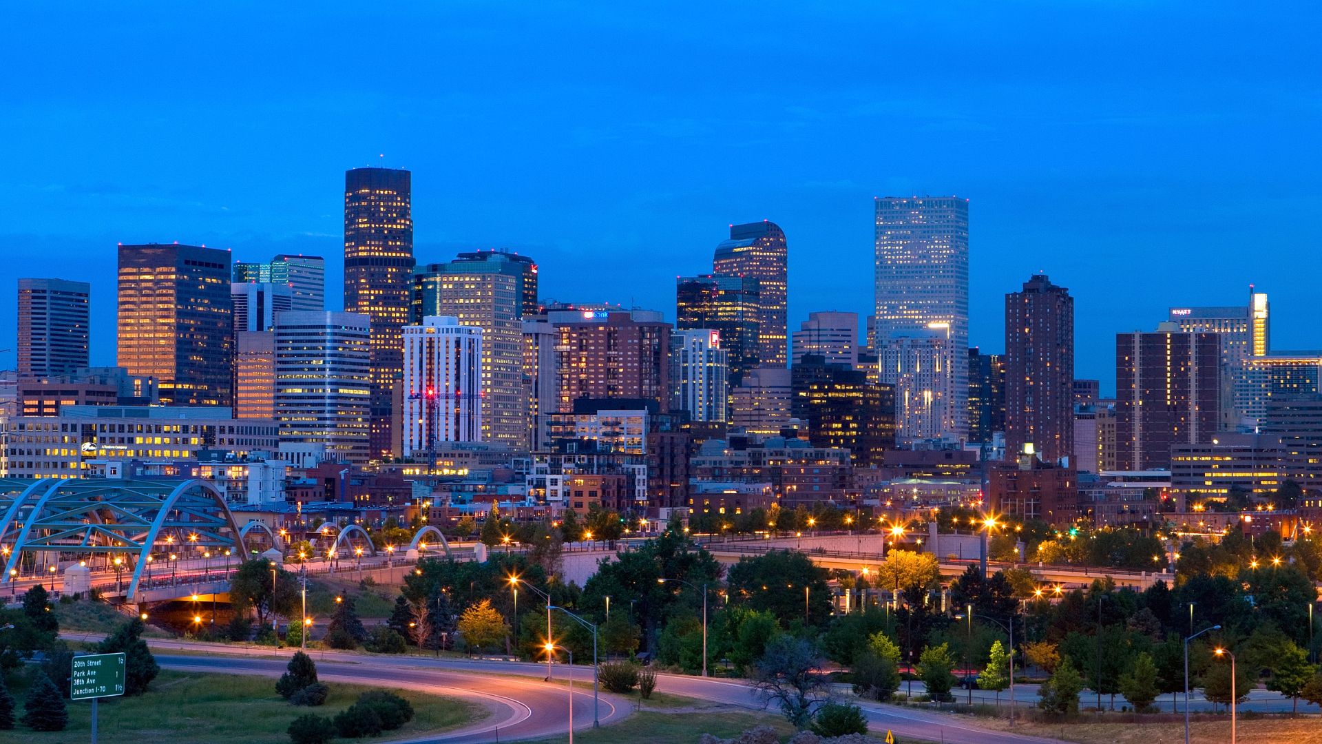 The illuminated downtown Denver skyline featuring numerous skyscrapers at dusk, with an arching bridge and tree-lined parkway in the foreground.