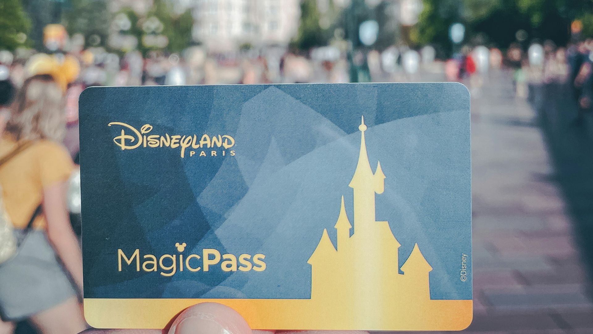 A hand holds a Disneyland Paris MagicPass in front of a blurred view of the theme park's entrance area.