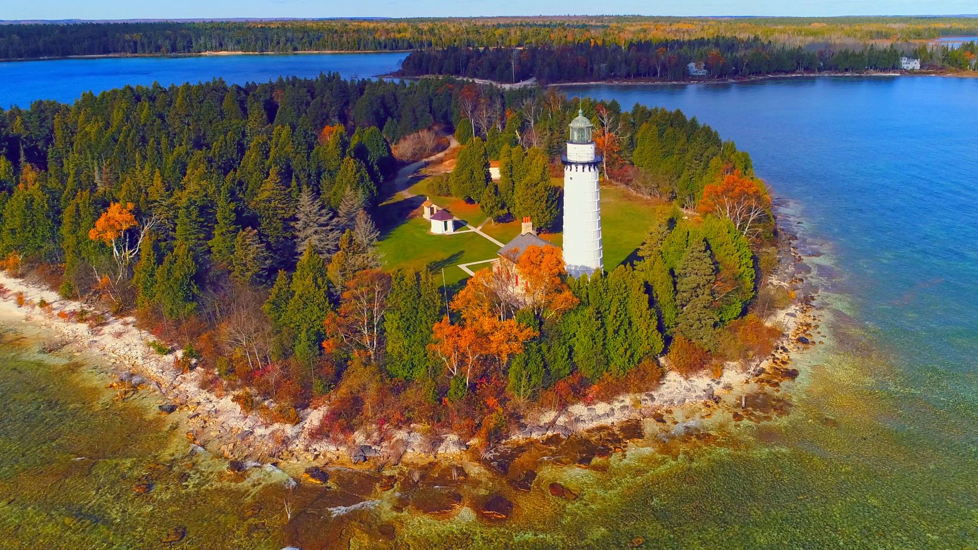 An aerial view of the Cana Island Lighthouse, a white tower on a small, tree-covered island, surrounded by a rocky shoreline and blue water, with a mainland forest in the distance showing autumn colors.