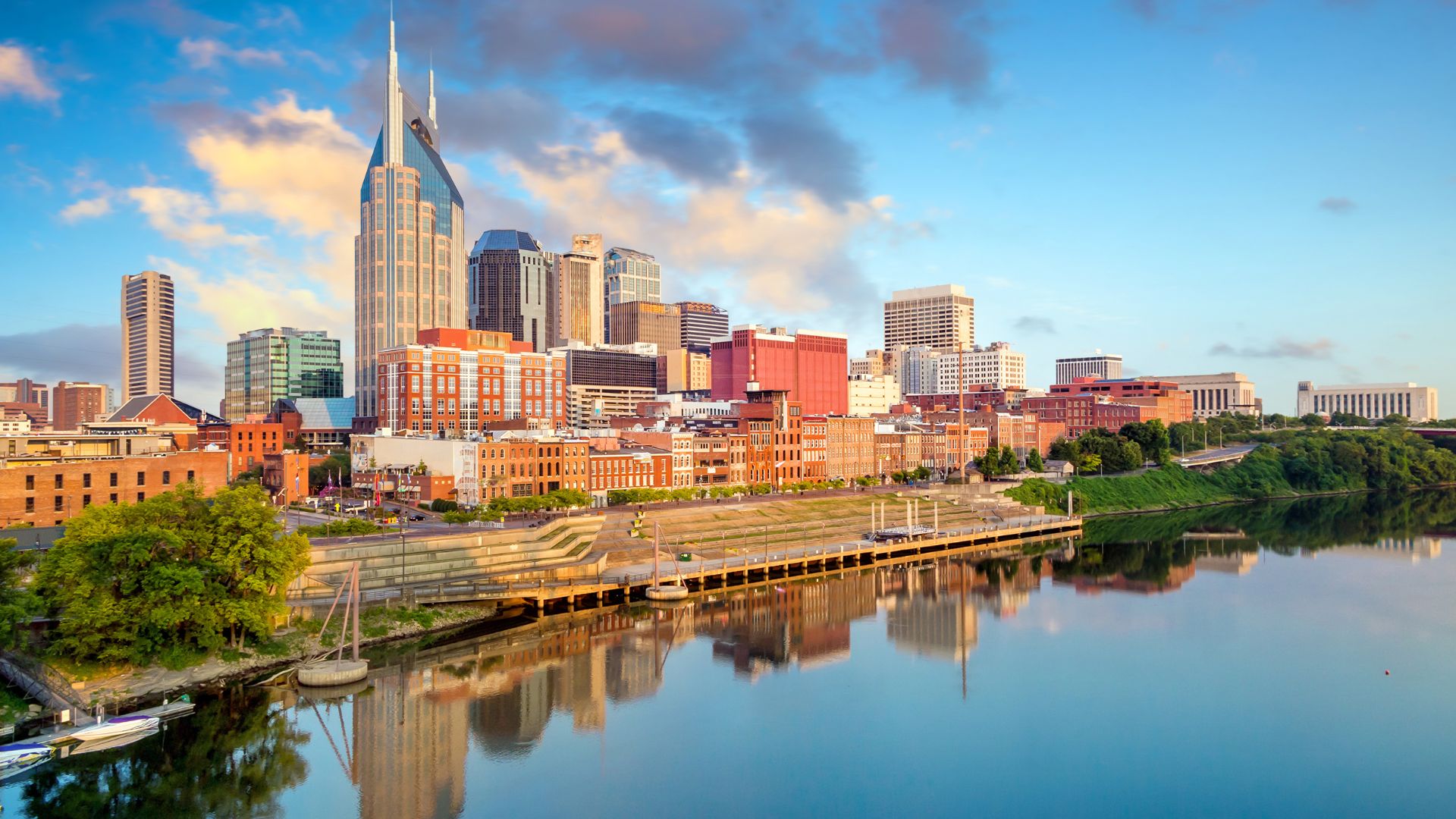 A daytime photo of the downtown Nashville, Tennessee skyline and the Cumberland River, with reflections of the buildings in the water. The tall "Batman Building" is prominent on the left.