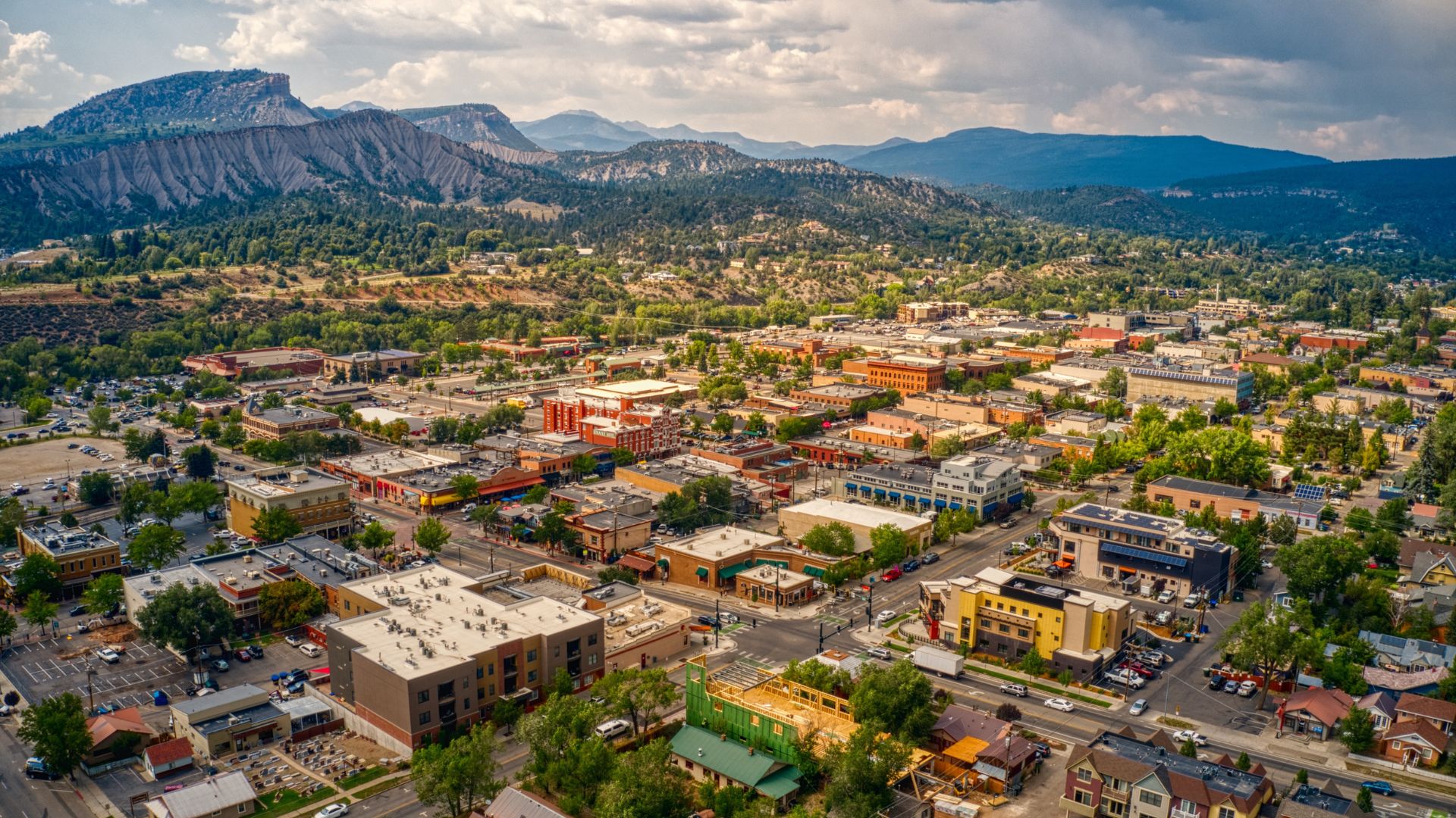 An aerial panoramic view of the downtown area of Durango, Colorado, with numerous buildings in a valley surrounded by dense forests and rugged mountains under a partly cloudy sky.