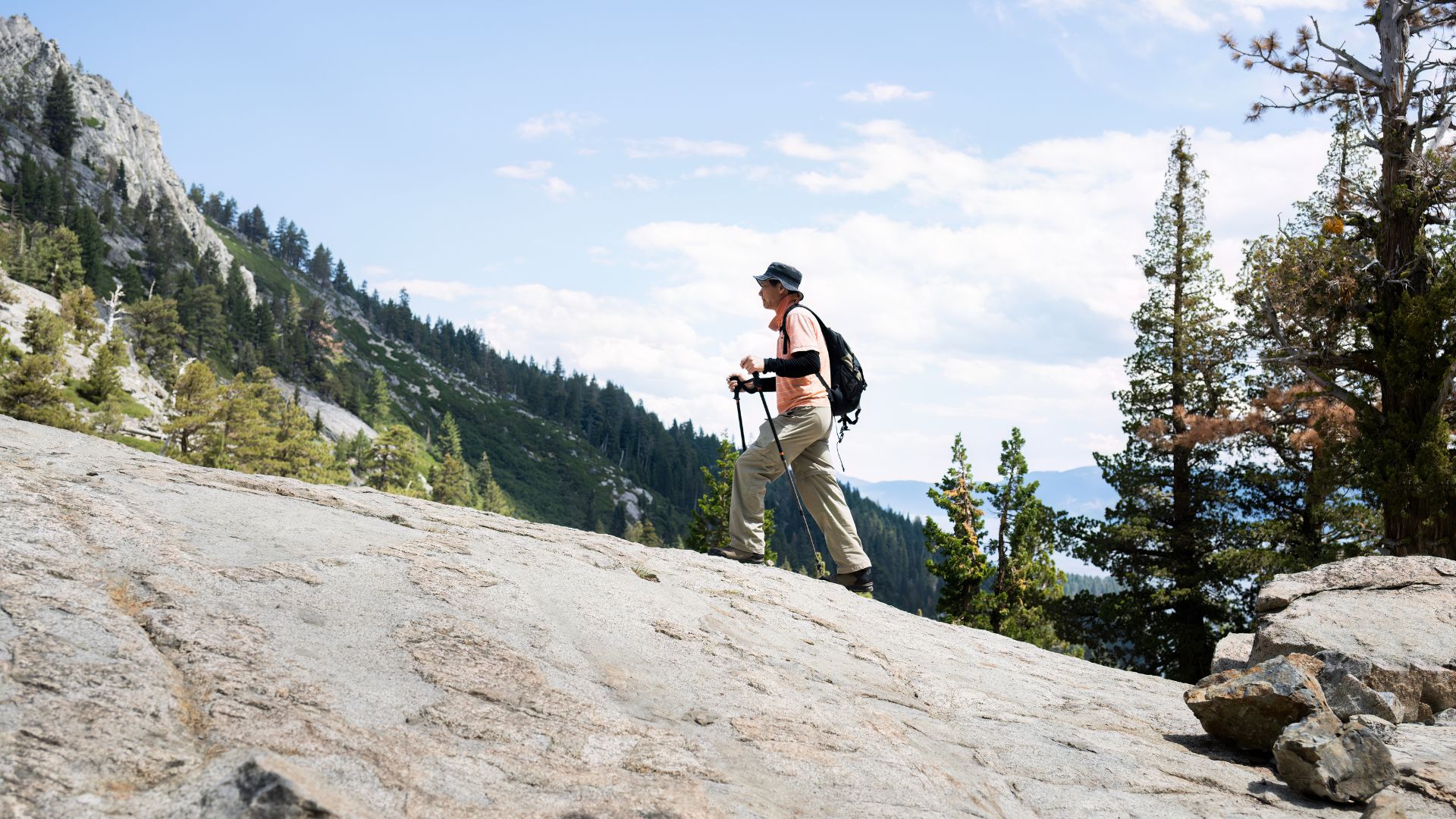 A hiker with a backpack and trekking poles walks along a large, sloping granite rock slab on the scenic Eagle Lake Trail in the Lake Tahoe wilderness, with dense green pine trees and distant mountains under a blue sky.