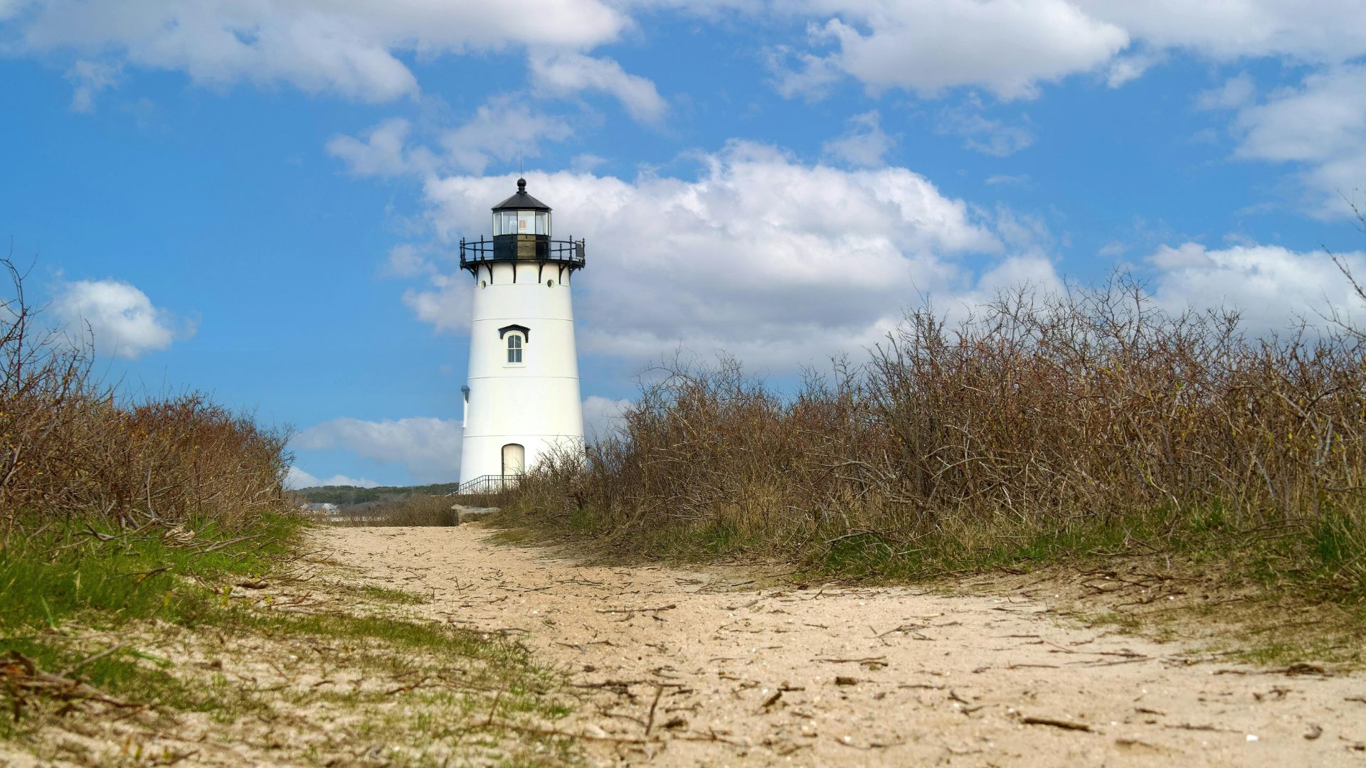 A white conical lighthouse with a black lantern room sits at the end of a sandy path, backed by dunes and scrub, under a blue sky with white clouds.