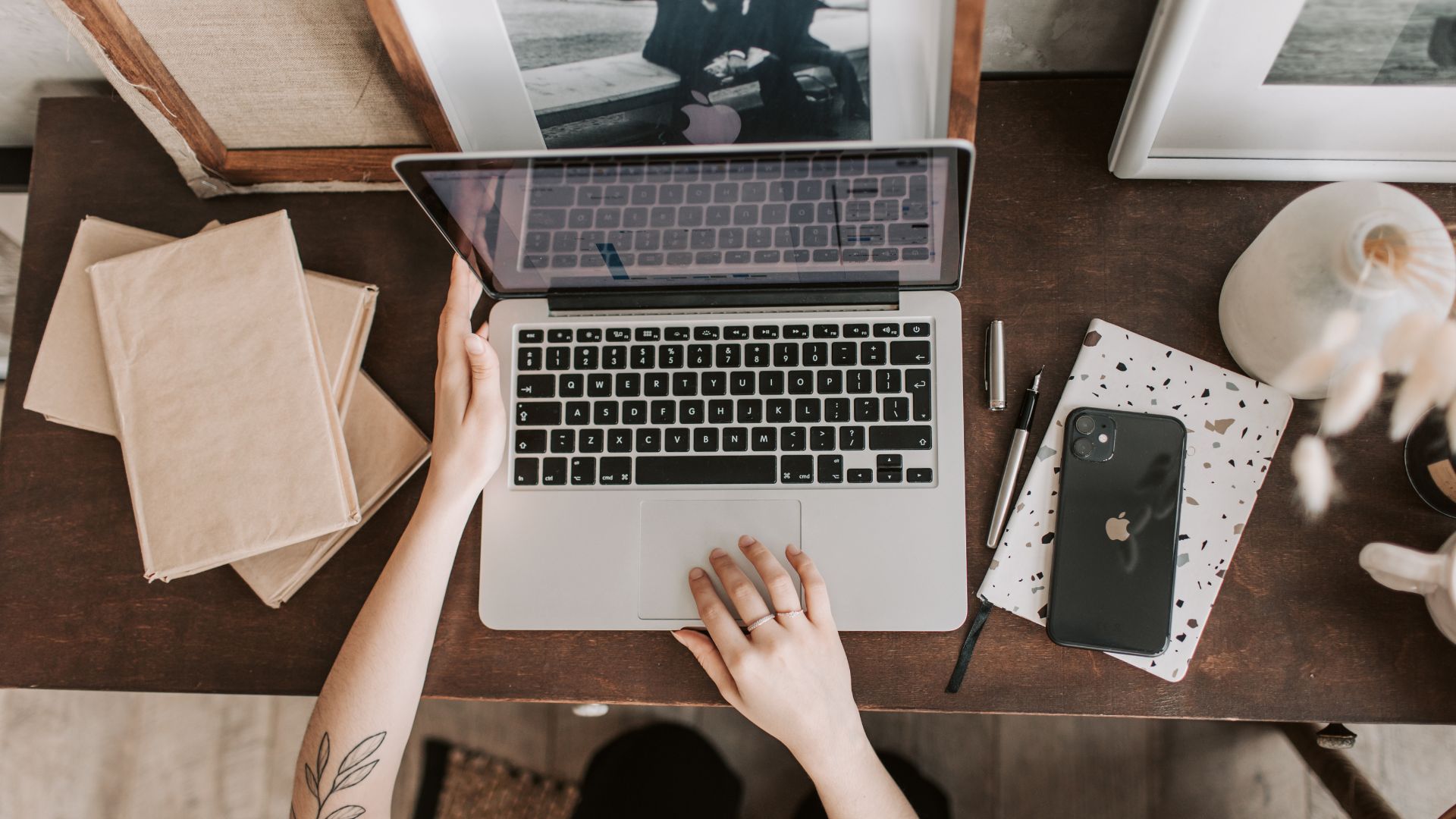 A top-down view of a person's hands on a silver laptop computer on a wooden desk, with a black smartphone, brown paper envelopes, and a patterned notebook nearby.