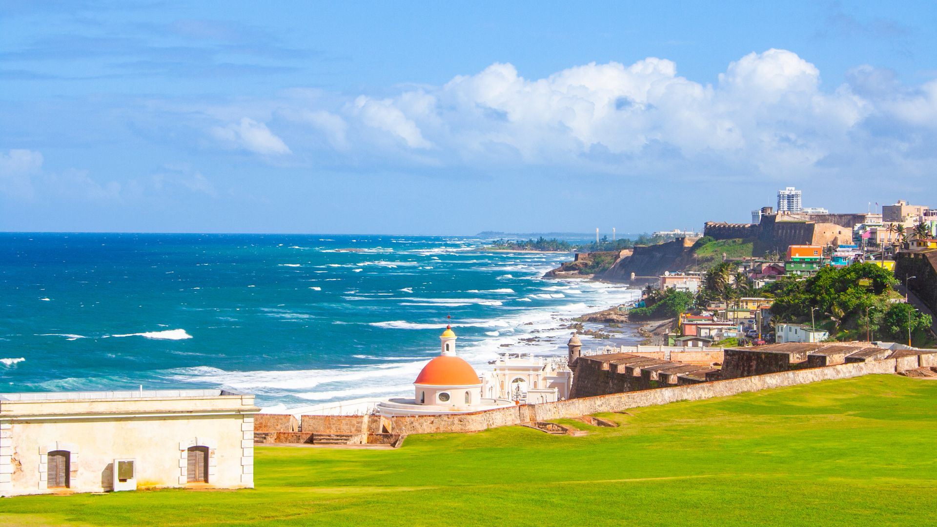 Panoramic view of the historic Castillo San Felipe del Morro fortress and the Atlantic Ocean coastline in San Juan, Puerto Rico, with a large green lawn in the foreground.