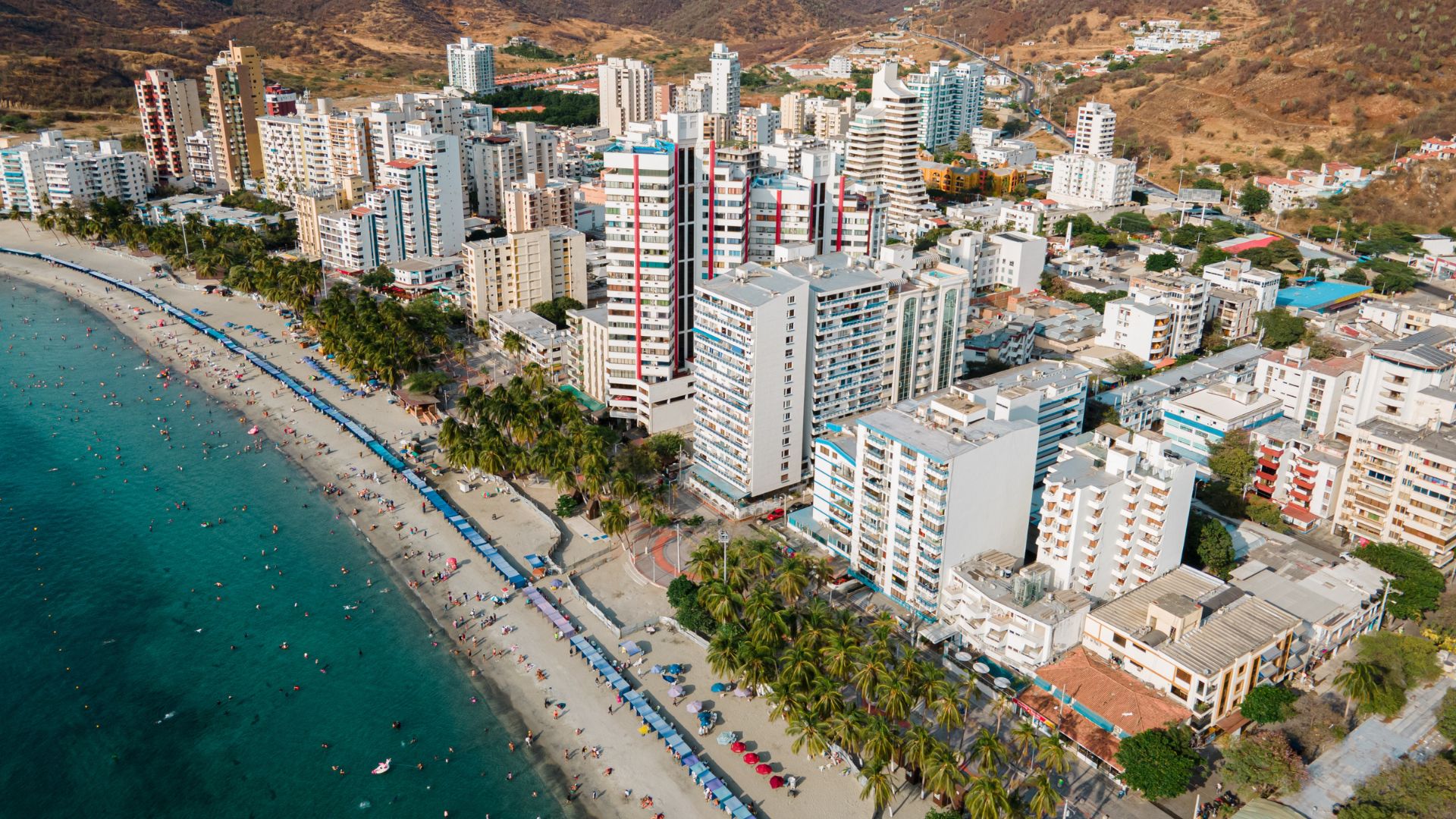An aerial view of the resort area of El Rodadero in Santa Marta, Colombia, showing a long, busy beach lined with high-rise hotels and buildings, with dry hills in the background.