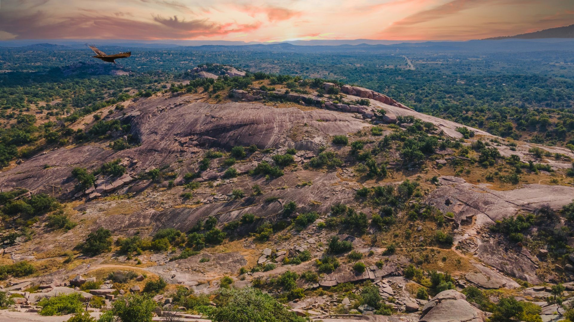 A large pink granite dome rises from the green, rolling hills of the Texas Hill Country under a vibrant, colorful sunset sky with a small airplane flying in the distance.