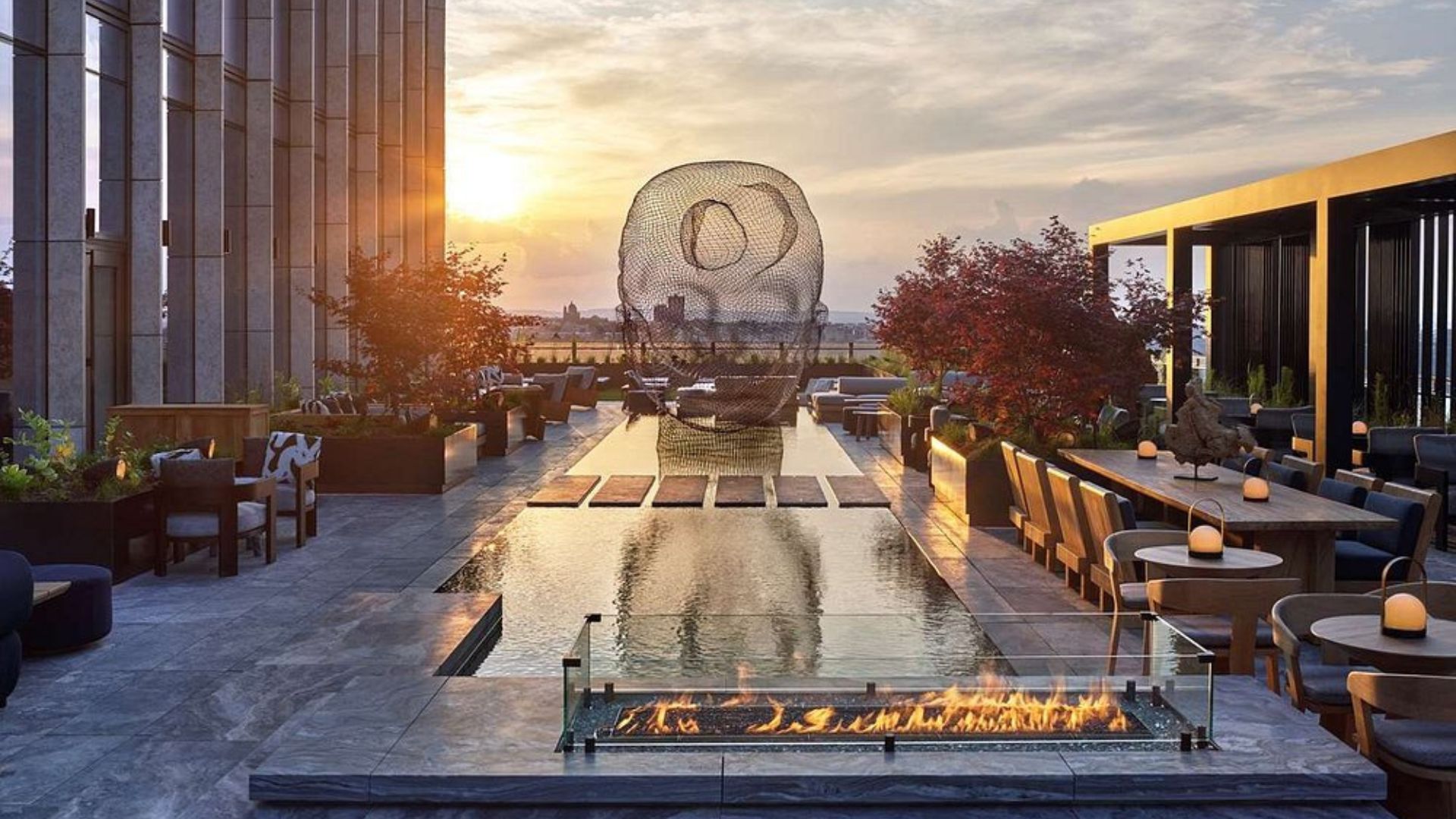 An expansive outdoor rooftop terrace at sunset, featuring a reflecting pool, a large wire-mesh sculpture of a head, dining tables, lounge seating, and a modern linear fire pit.