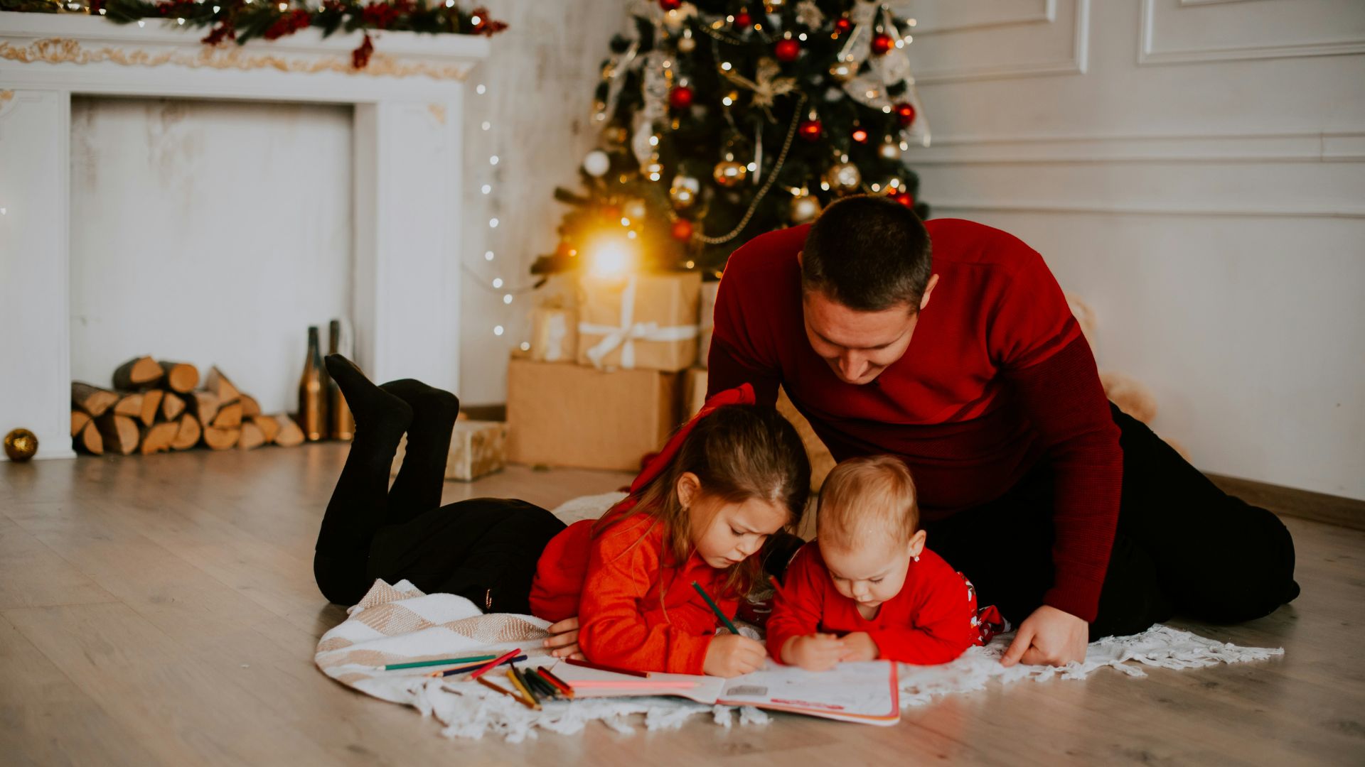 A father and two young children sit on a rug in front of a fireplace and Christmas tree, coloring in a book.