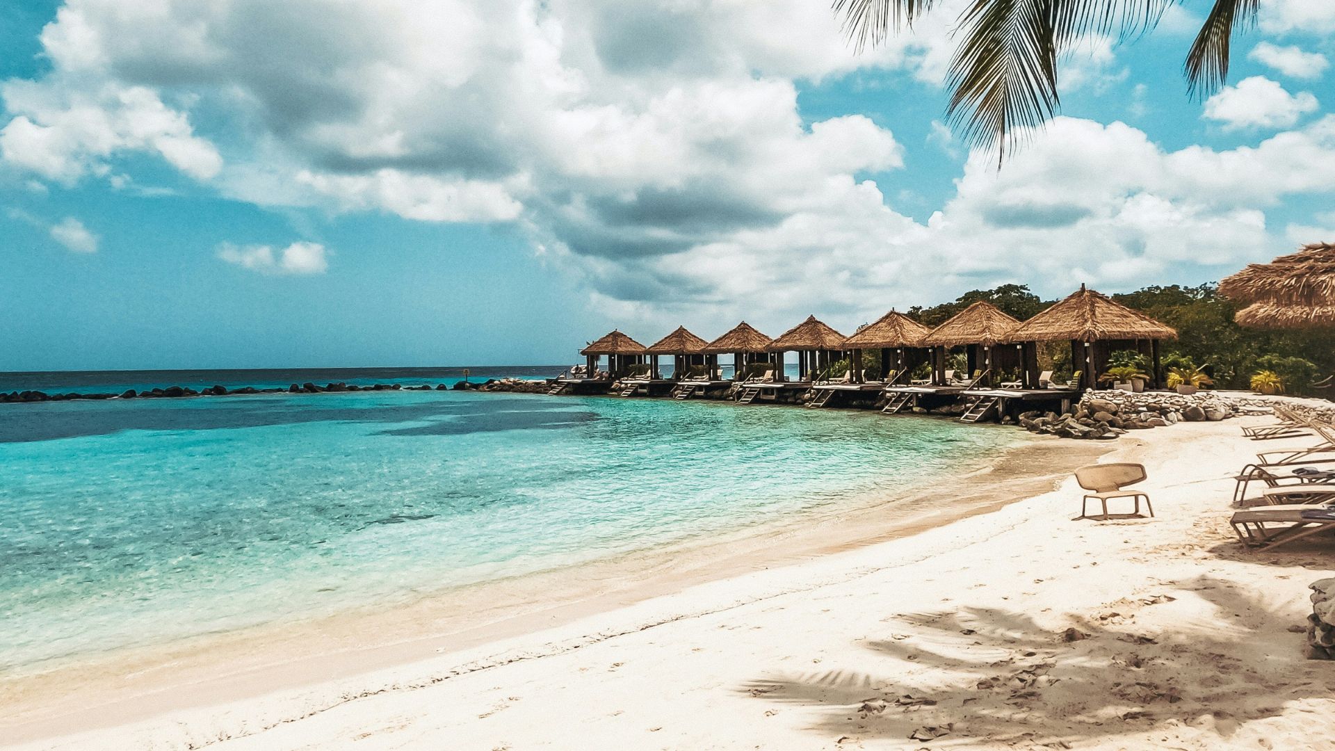 A panoramic view of Flamingo Beach on Renaissance Island, Aruba, showing a white sand beach with several thatched-roof huts and calm turquoise water.