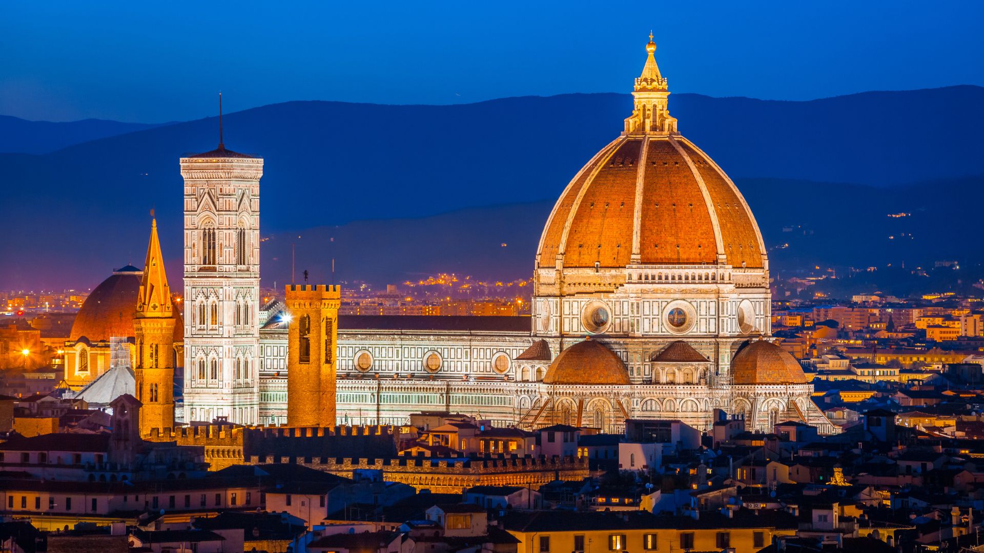 A nighttime panoramic view of the historic city of Florence featuring the illuminated, red-domed cathedral and its tall bell tower against a deep blue sky with mountains in the background.