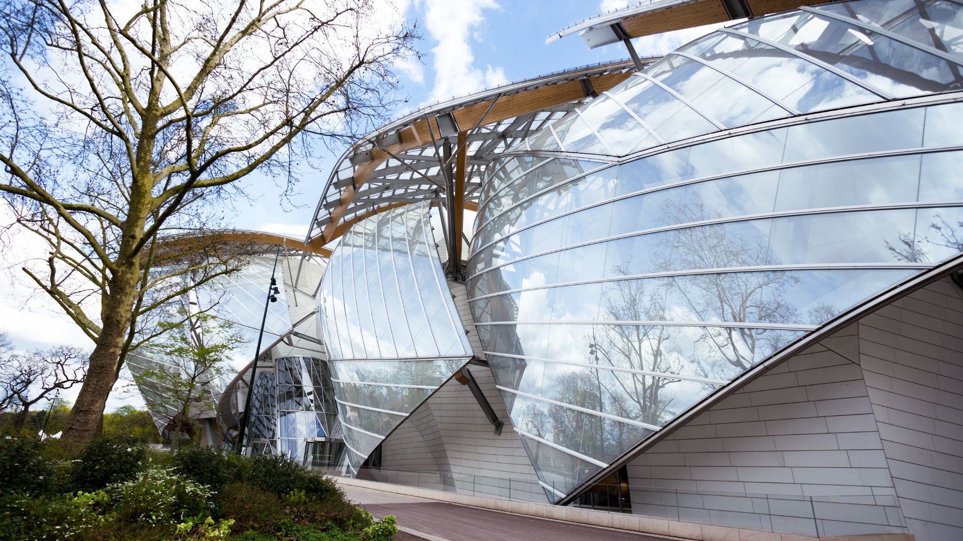 A futuristic museum building with large, curving glass "sails" reflecting trees, set next to a walkway and garden.