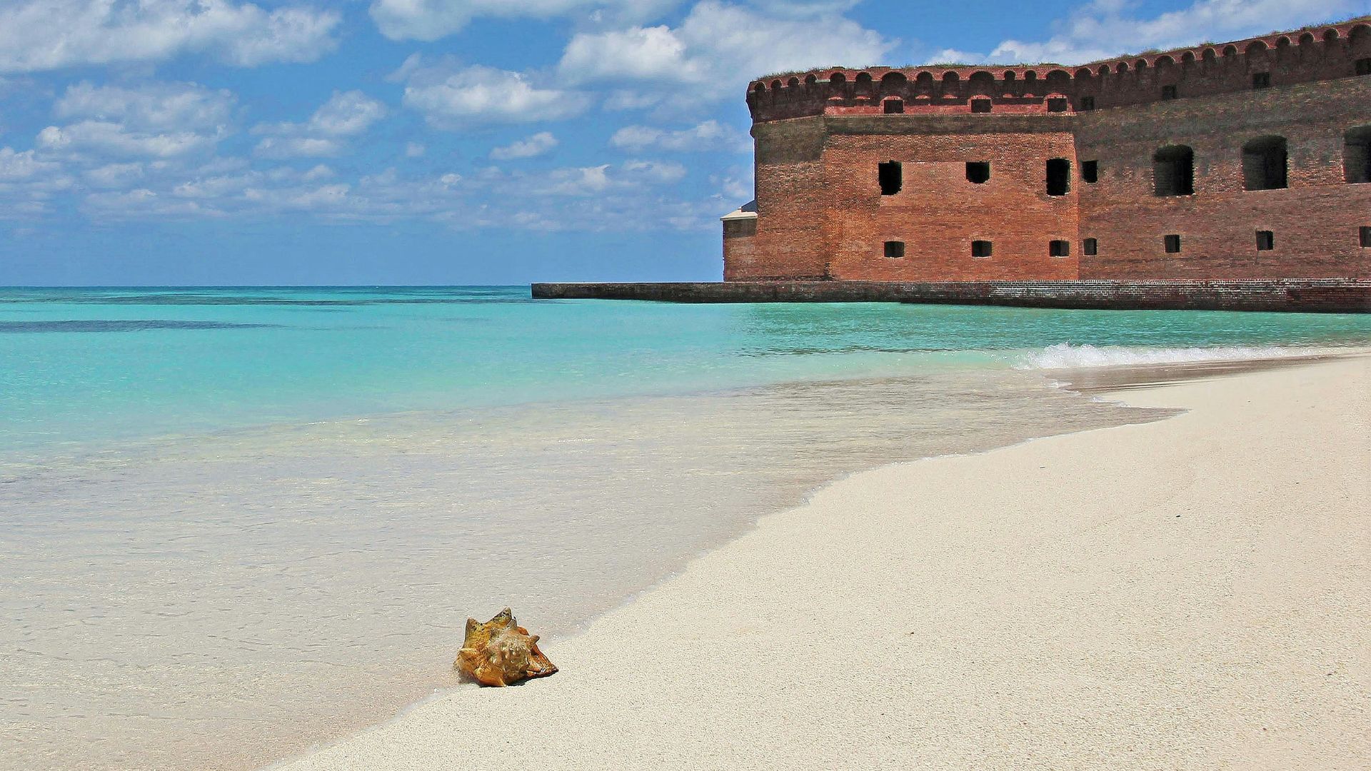 A large, red-brick 19th-century fort stands next to a white sandy beach and clear turquoise water, with a conch shell in the foreground.