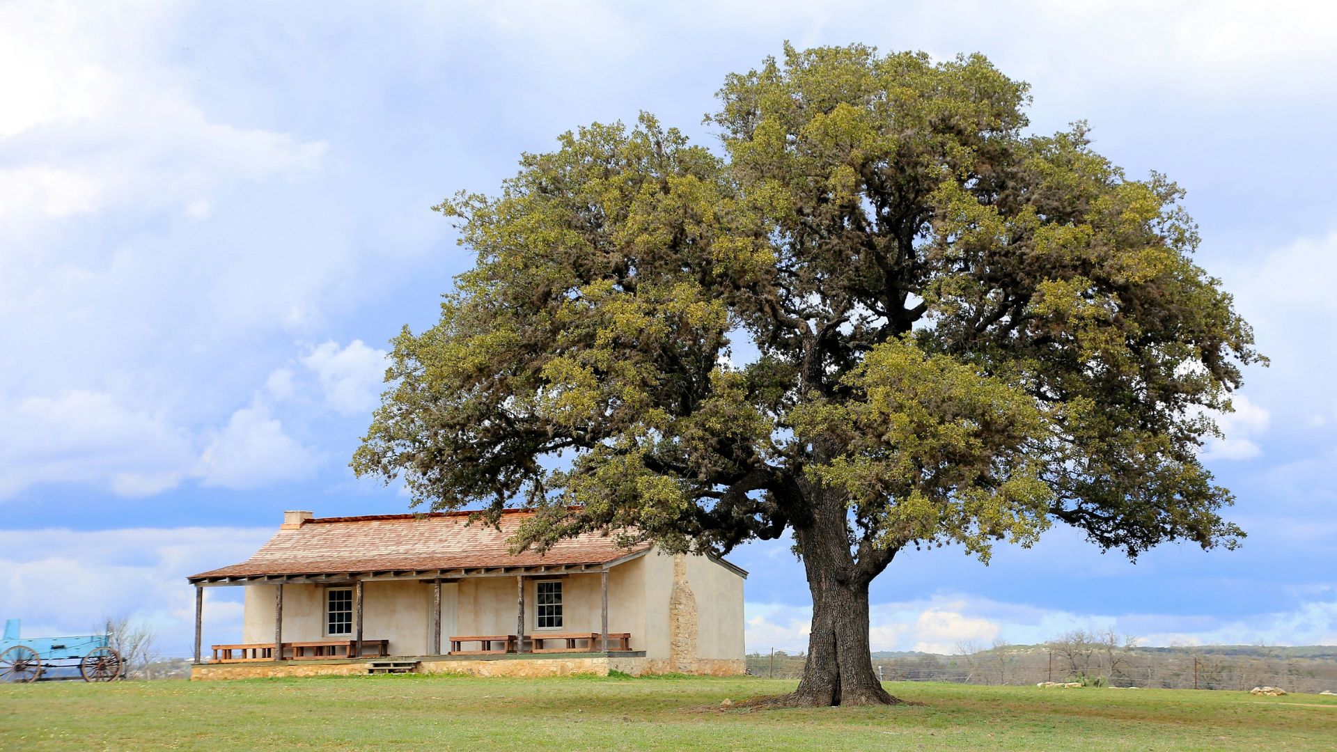 A historic, single-story adobe building with a covered porch and a red-tile roof sits in a green field next to a very large, mature oak tree under a partly cloudy sky.