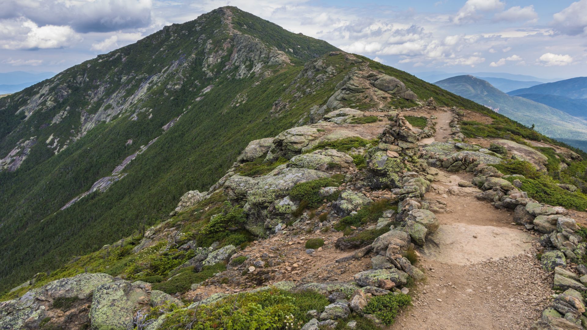 A rugged, rocky hiking trail follows an exposed mountain ridge with low green vegetation and a low stone wall, leading toward a steep, tree-covered mountain peak in the distance under a blue and cloudy sky.