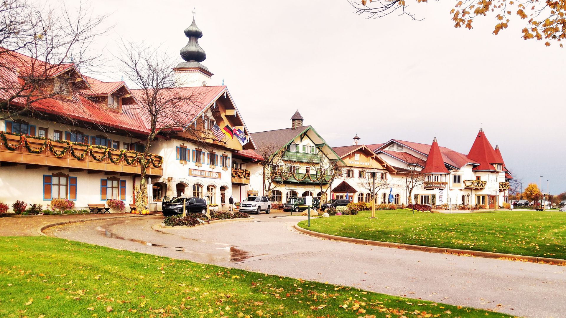 A row of traditional German-style buildings with white facades, red-tiled roofs, and dark wood trim lines a paved driveway next to a green lawn, under an overcast sky with hints of autumn foliage in the upper right.