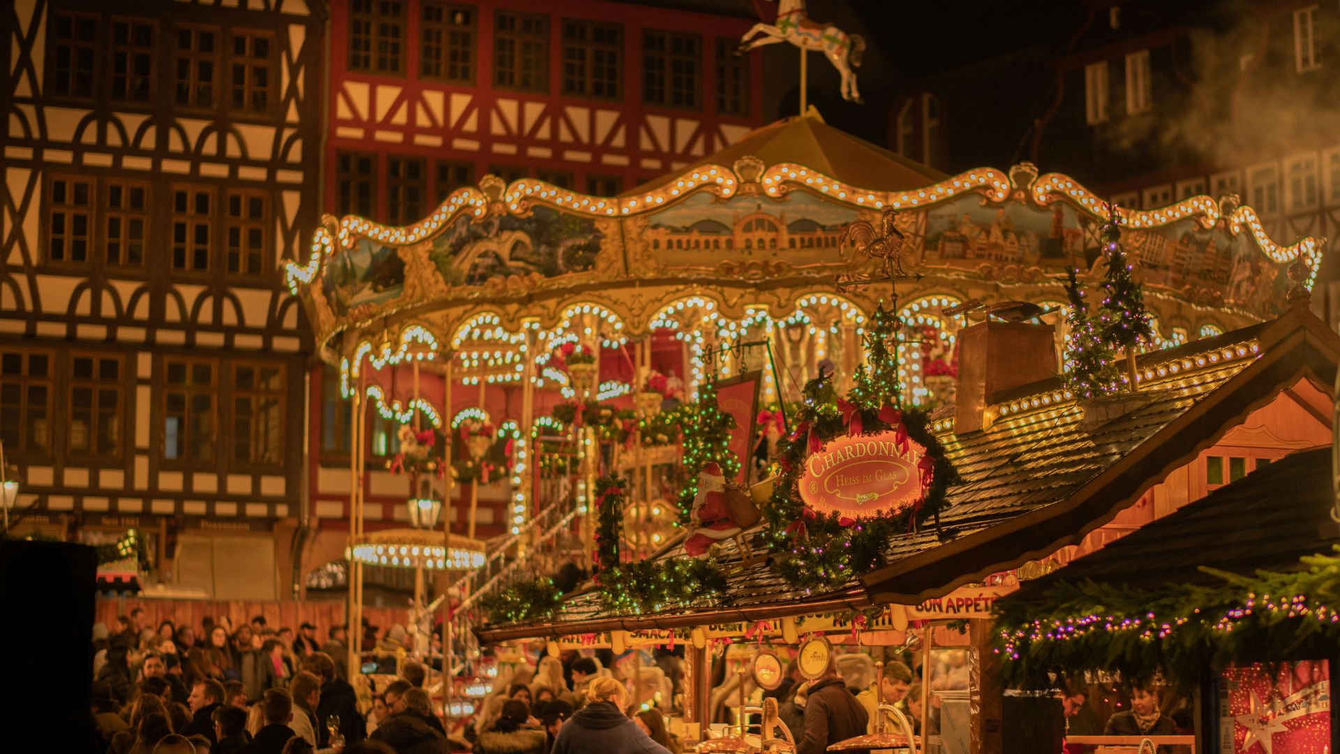 A vibrant night scene of the Frankfurt Christmas Market featuring a lit-up carousel, festive stalls, and traditional half-timbered buildings.
