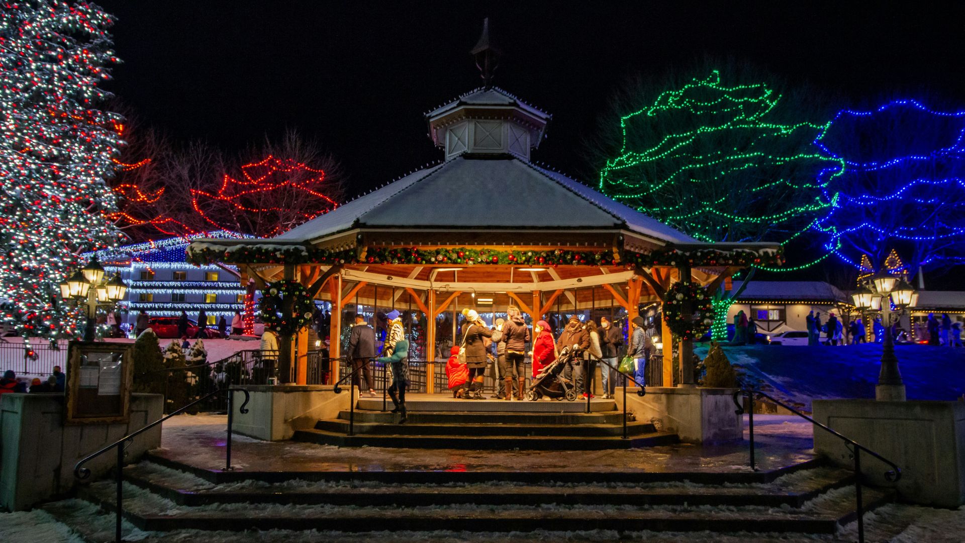 A snowy, nighttime view of a wooden gazebo in a park, brightly lit with Christmas lights. People are gathered inside the gazebo, and large trees in the background are covered in red, green, and blue lights.
