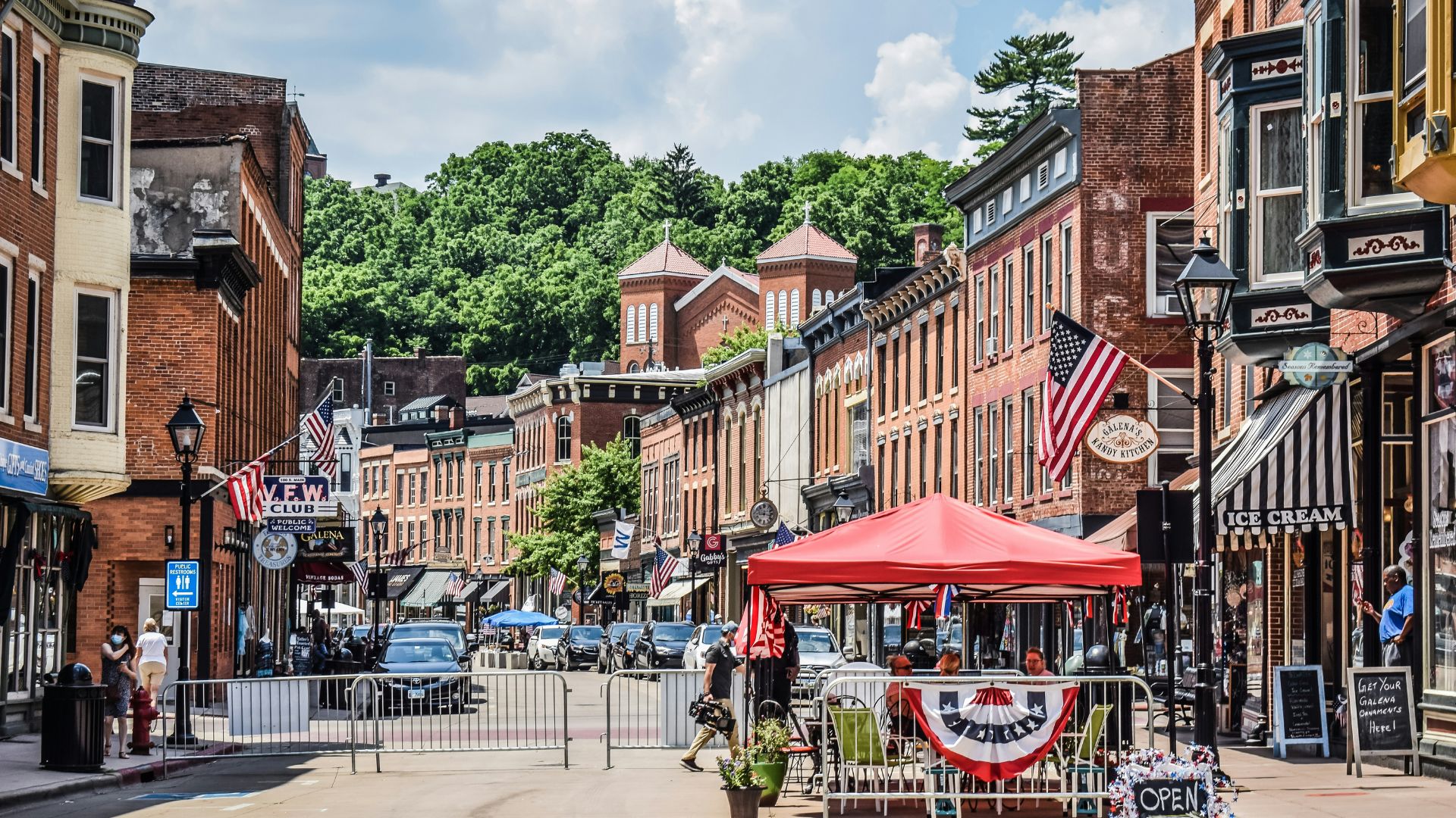 A wide-angle view of the historic Main Street in Galena, Illinois, lined with red-brick buildings and American flags, with a red tent and street closure barriers in the foreground and a steep, tree-covered hill in the background.