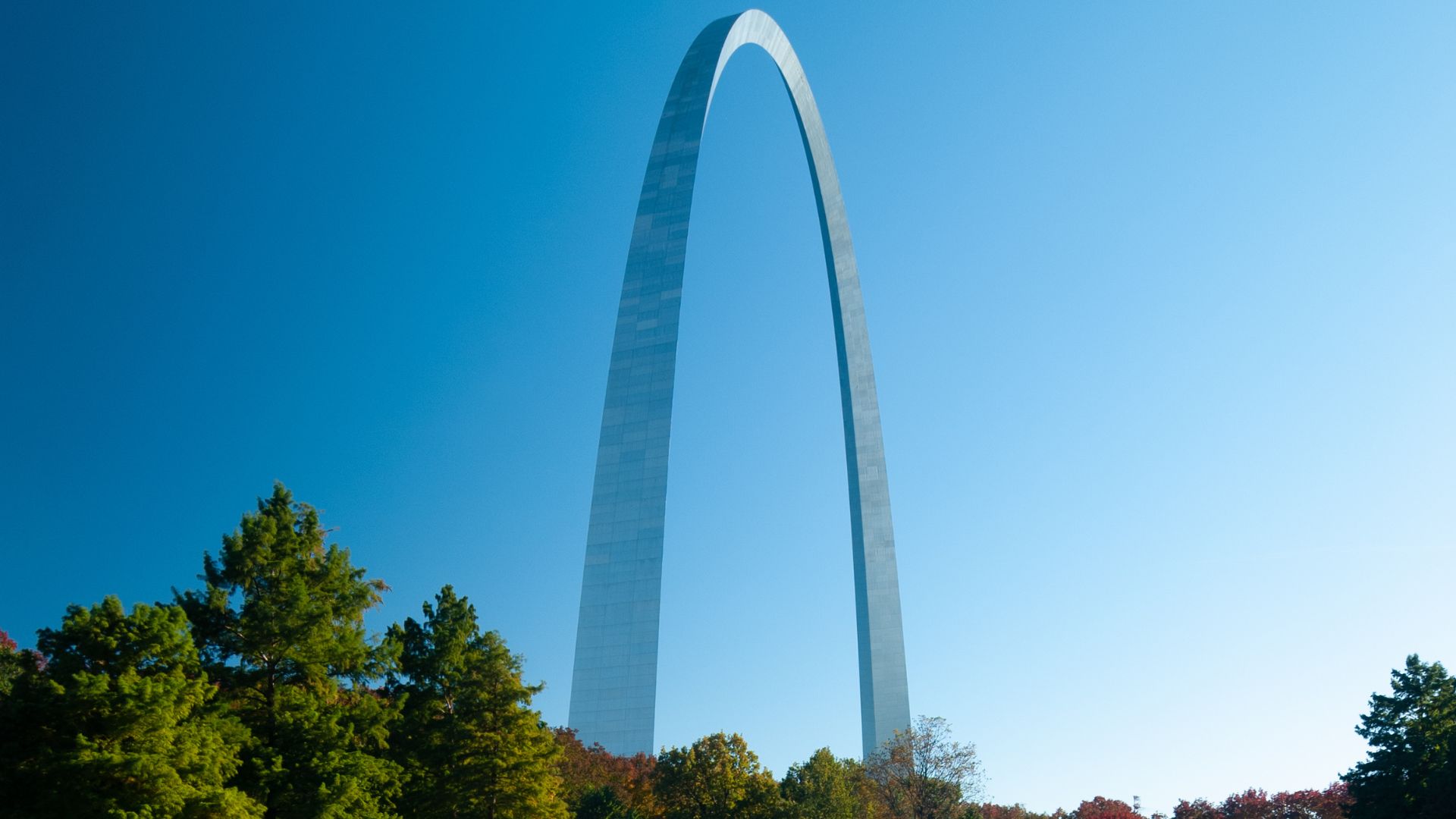 A massive, gleaming stainless steel arch rises dramatically against a bright blue sky, framed by green trees with hints of autumn foliage in the foreground.