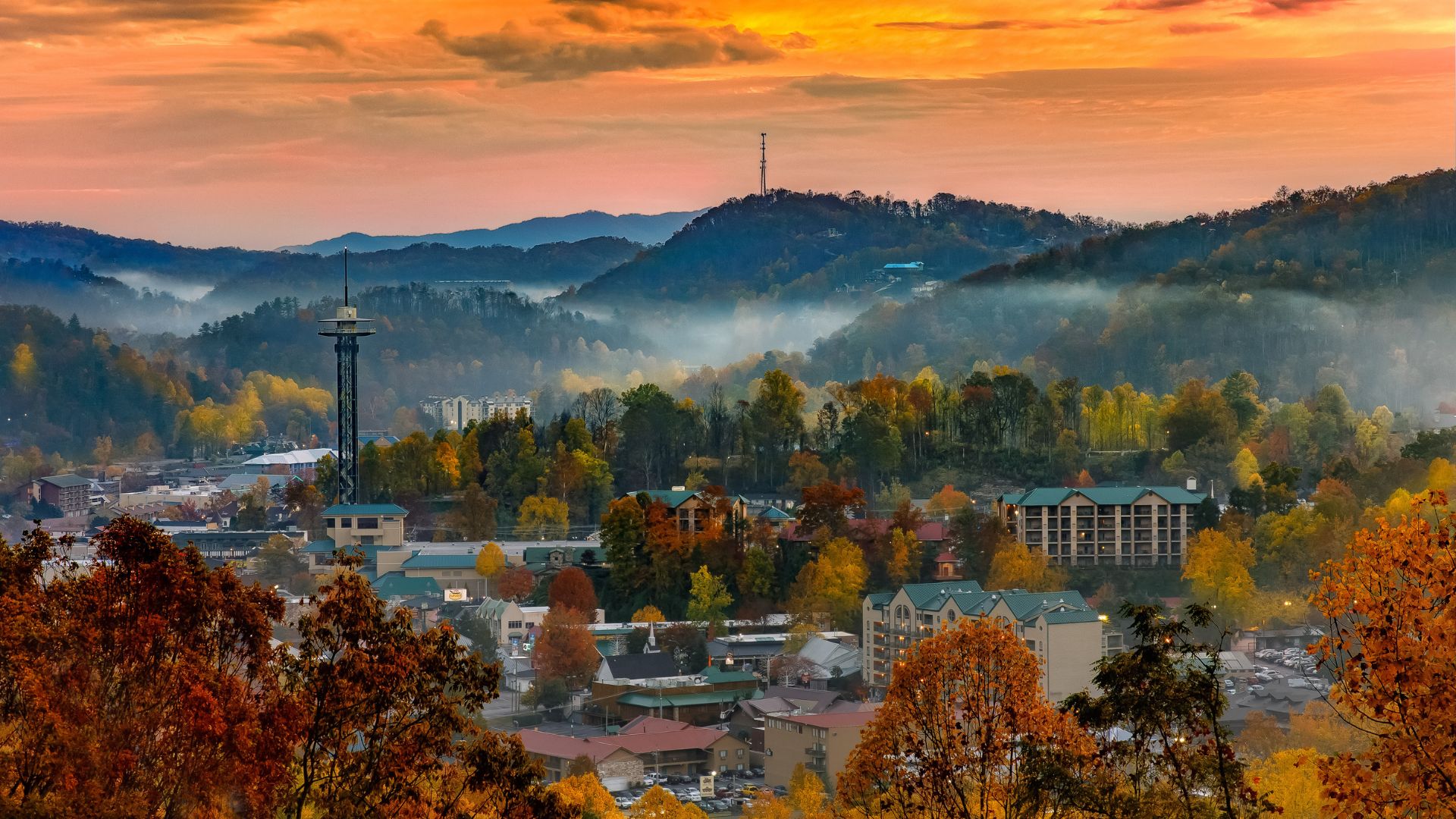 An aerial view of the town of Gatlinburg, Tennessee, in the autumn, featuring a prominent observation tower and buildings surrounded by trees with vibrant fall colors and mist rolling over the mountains in the background.