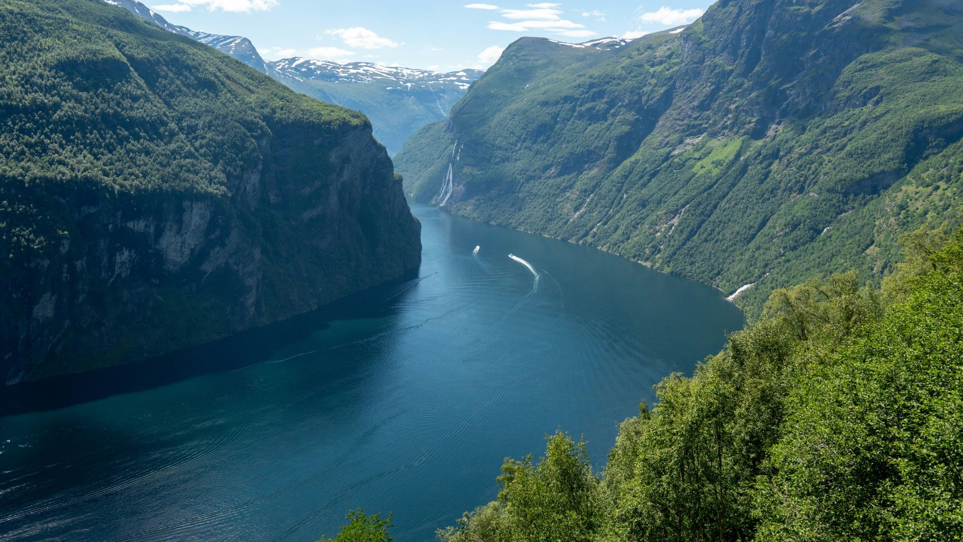 A view of the Geirangerfjord in Norway from an elevated wooden cabin deck, with steep, green mountainsides surrounding the blue water where a small white boat is cruising.