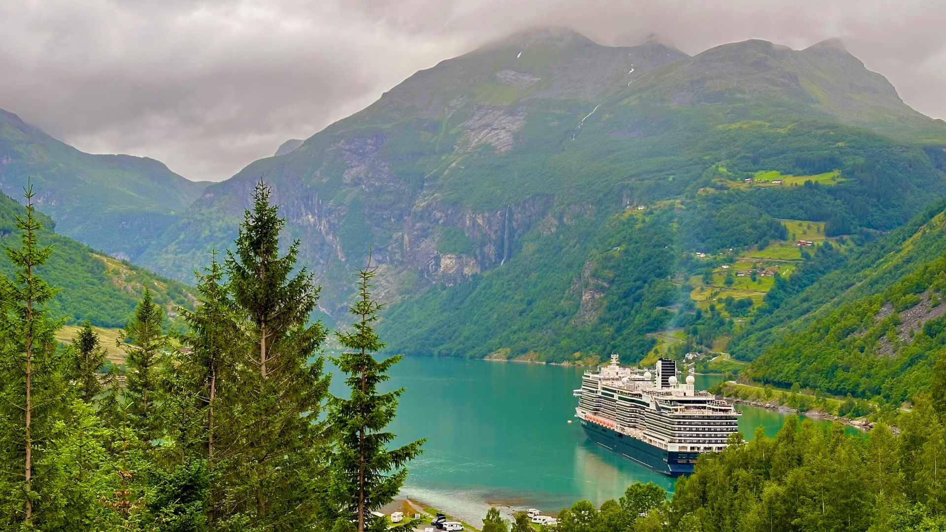 A large cruise ship sailing in a narrow, green Norwegian fjord surrounded by towering, lush green mountains under a cloudy sky.