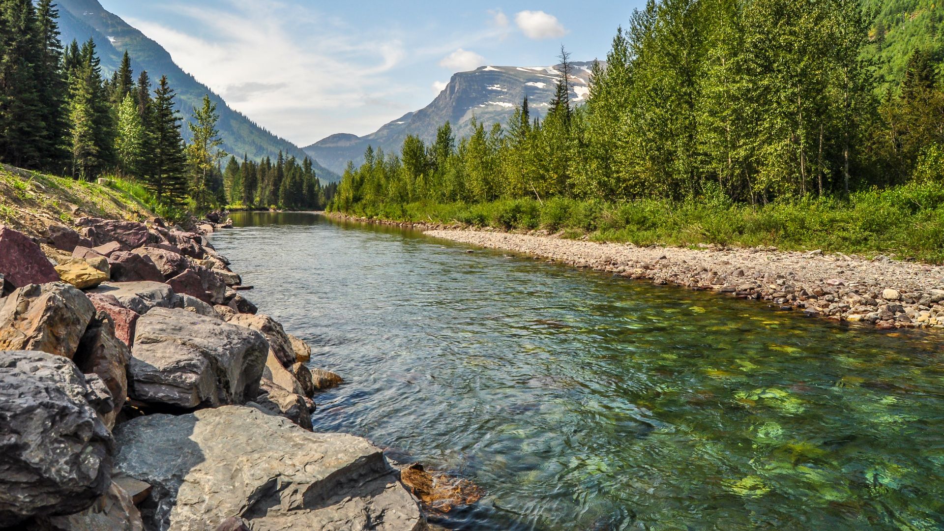 A clear, rocky mountain river flows from the foreground into a valley surrounded by steep, pine-forested mountains under a blue sky with white clouds.