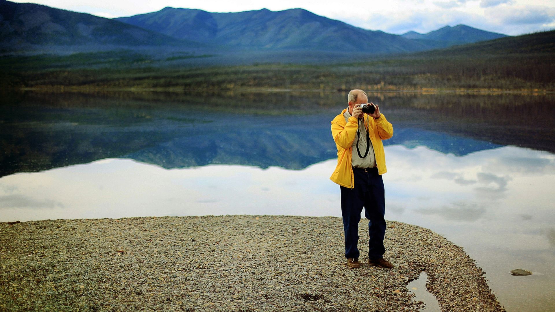 A man in a yellow jacket stands on the gravel shore of a calm lake, photographing a perfect reflection of mountains and trees in the water under a cloudy sky.