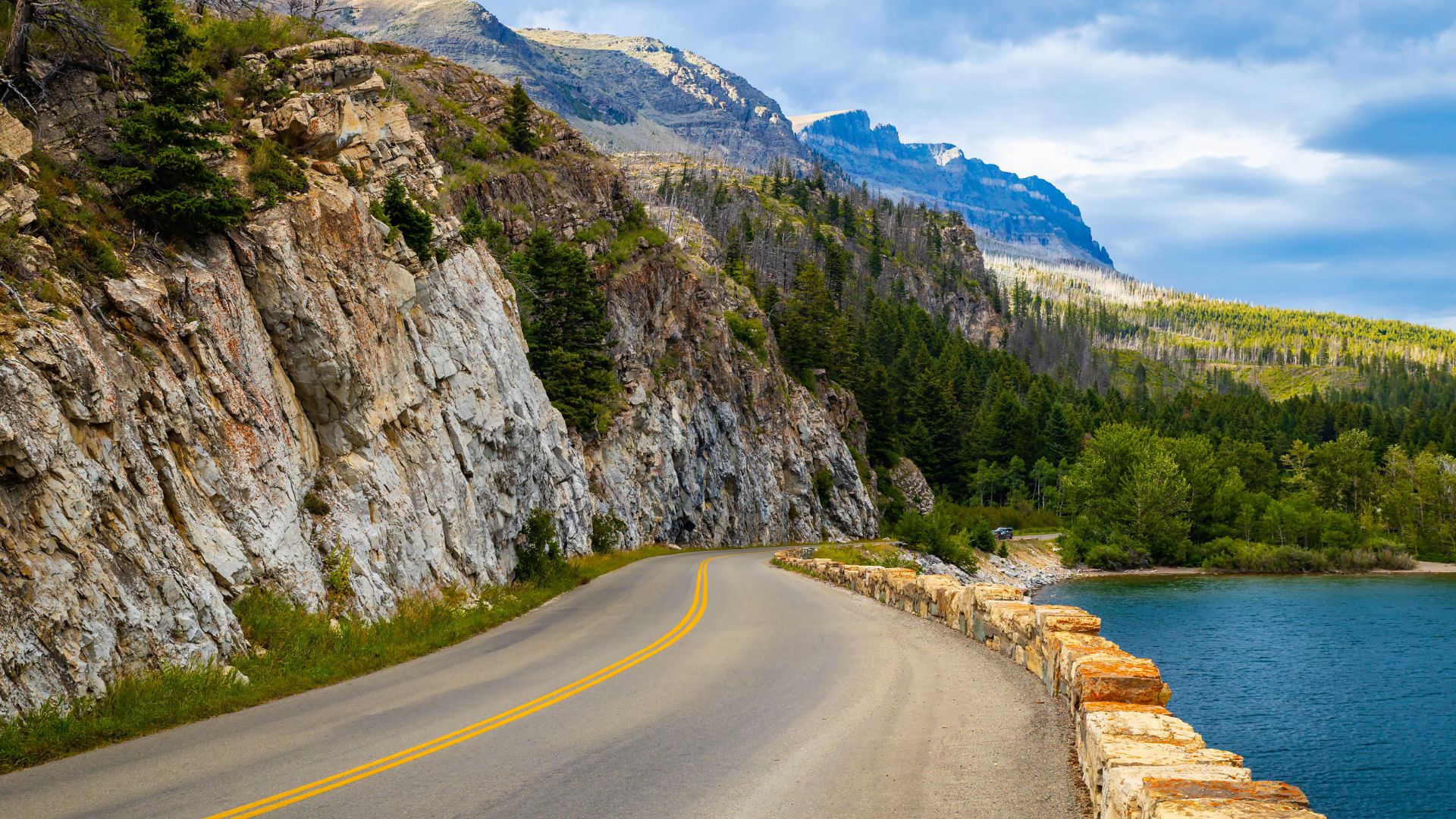 A winding mountain road with a low stone retaining wall on the right runs alongside a blue lake, with steep, rocky cliffs rising on the left and large, forested mountains filling the background under a blue sky.