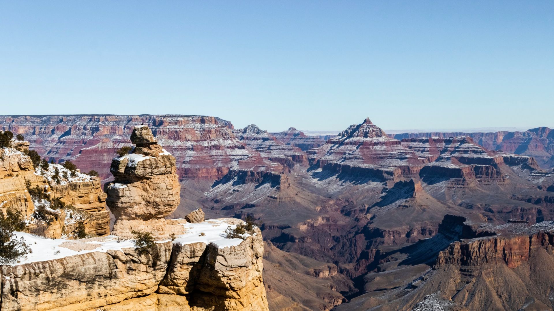 A panoramic, sunny view of the snow-dusted Grand Canyon in Arizona, featuring a prominent, duck-shaped rock formation in the foreground and layered canyon walls in the distance.