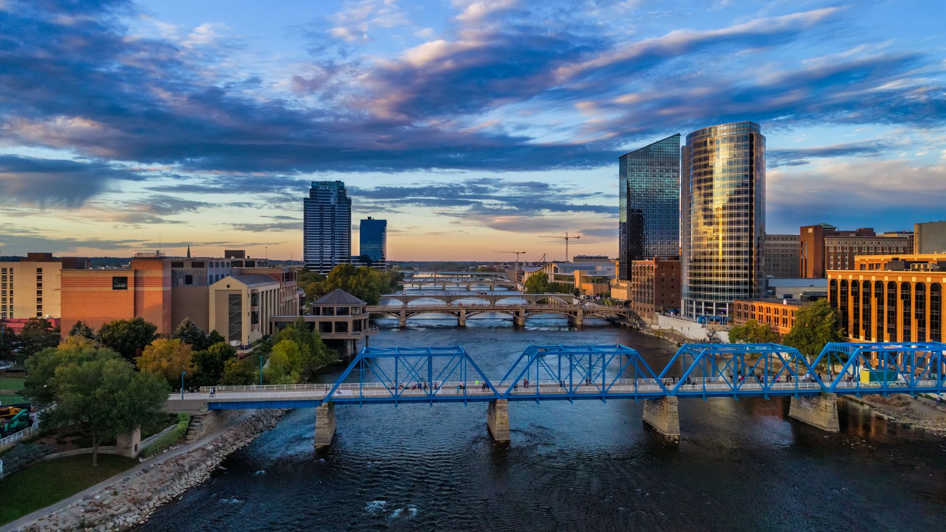 A photo of the Grand Rapids, Michigan skyline at sunset, with the iconic blue pedestrian truss bridge spanning the Grand River in the foreground.