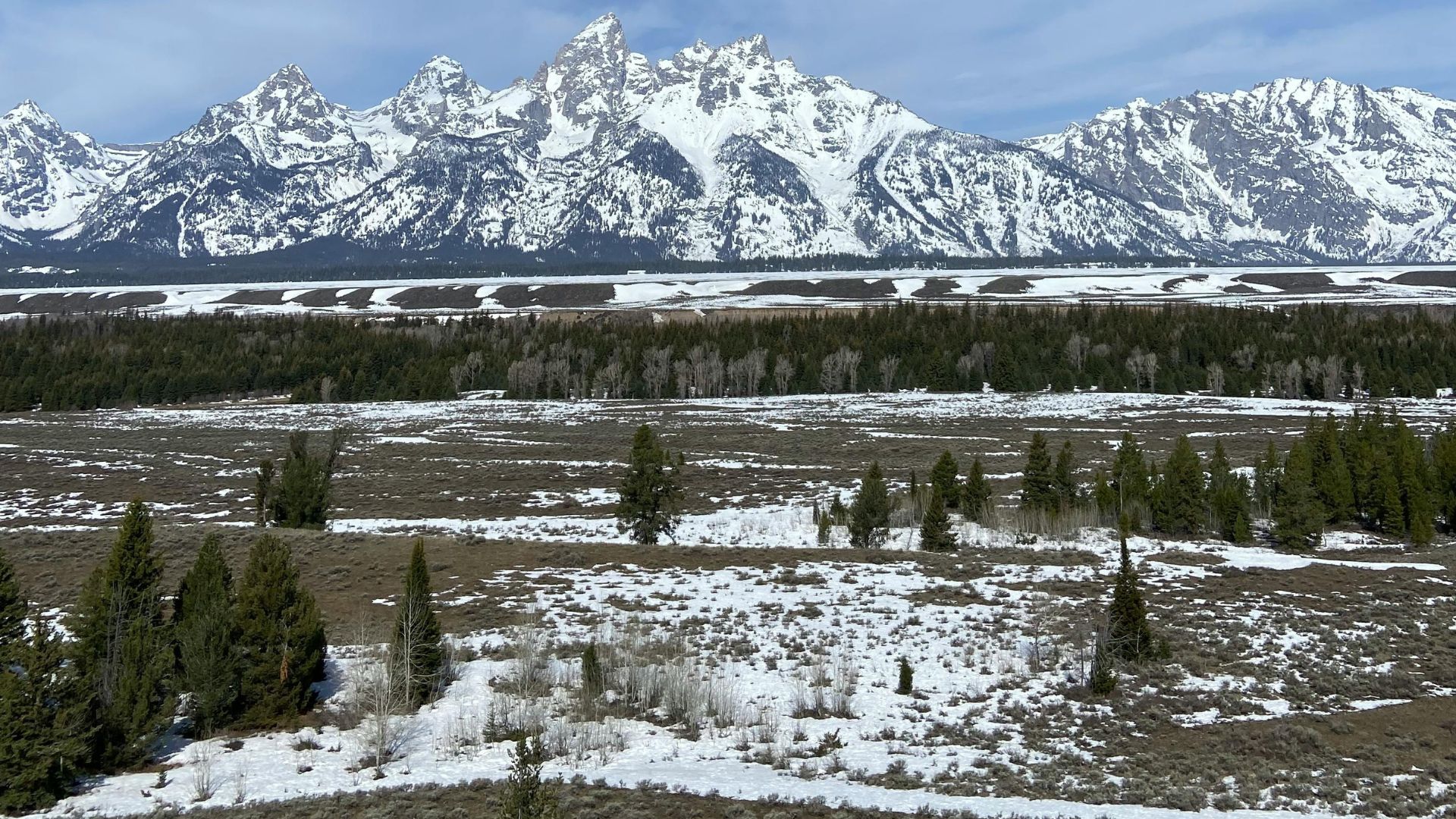 A panoramic view of the snow-capped Teton mountain range under a bright blue sky with white clouds.