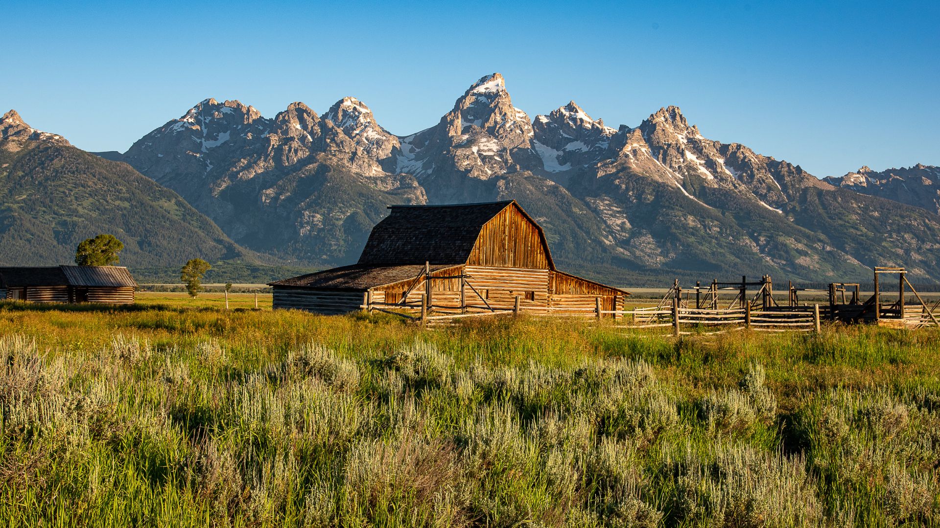 A wooden barn sits in a grassy field, with a rugged, snow-capped mountain range rising in the background under a clear blue sky.