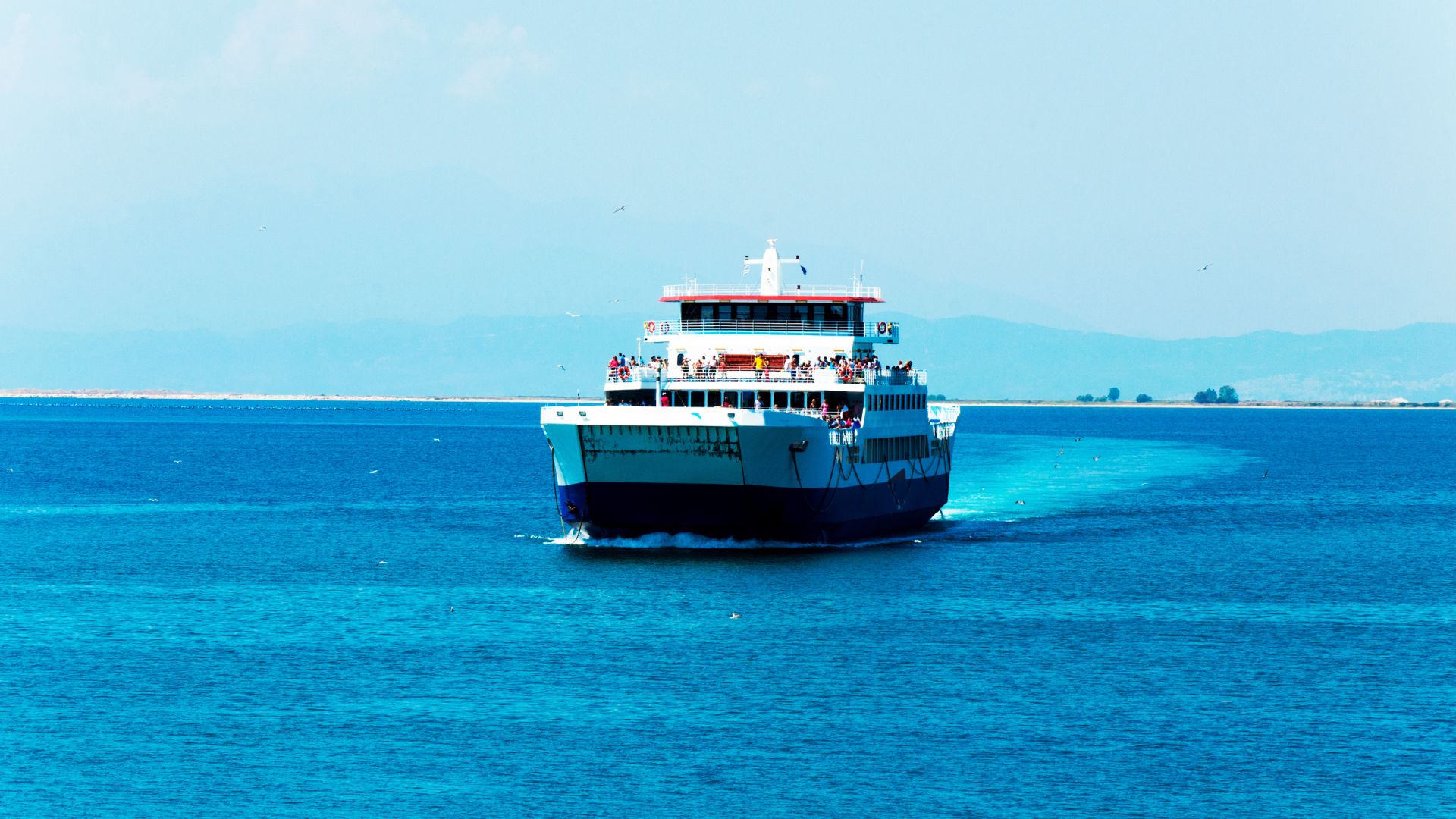 A medium-sized passenger and car ferry with a white and blue hull moves through bright turquoise blue water, leaving a white wake behind it, with low hills visible on the horizon.