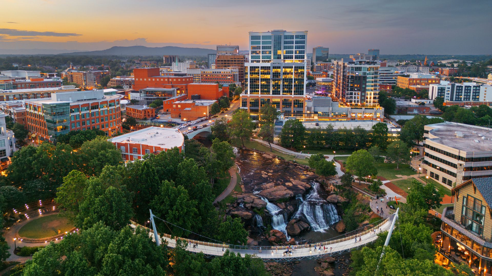 An aerial dusk photo of the Greenville, South Carolina skyline and a curved pedestrian suspension bridge over a series of waterfalls in a lush park.