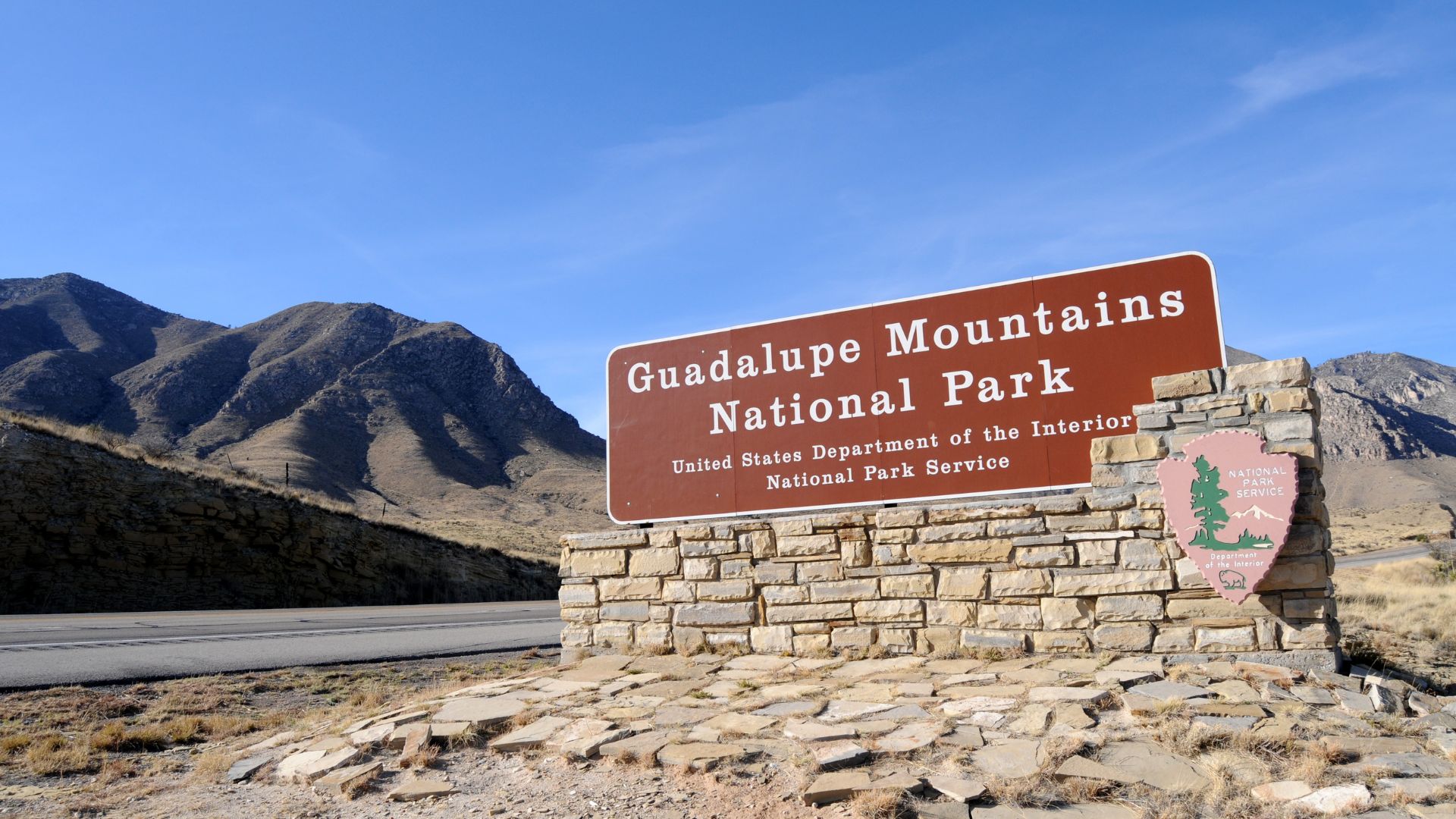 A large, brown national park entrance sign made of rock and wood stands beside a paved road with steep, arid mountains rising sharply in the background under a clear blue sky.