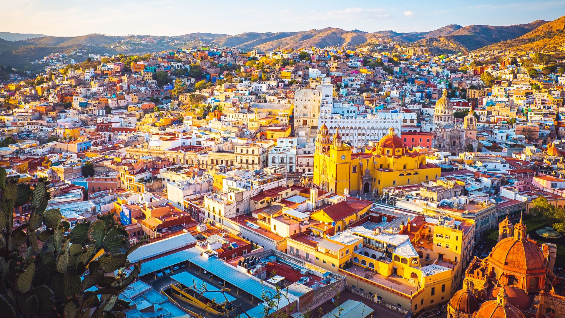 A panoramic, elevated view of the historic city of Guanajuato, Mexico, showing a dense cluster of vibrantly painted, multi-colored buildings nestled in a mountain valley under a bright sky with golden light.