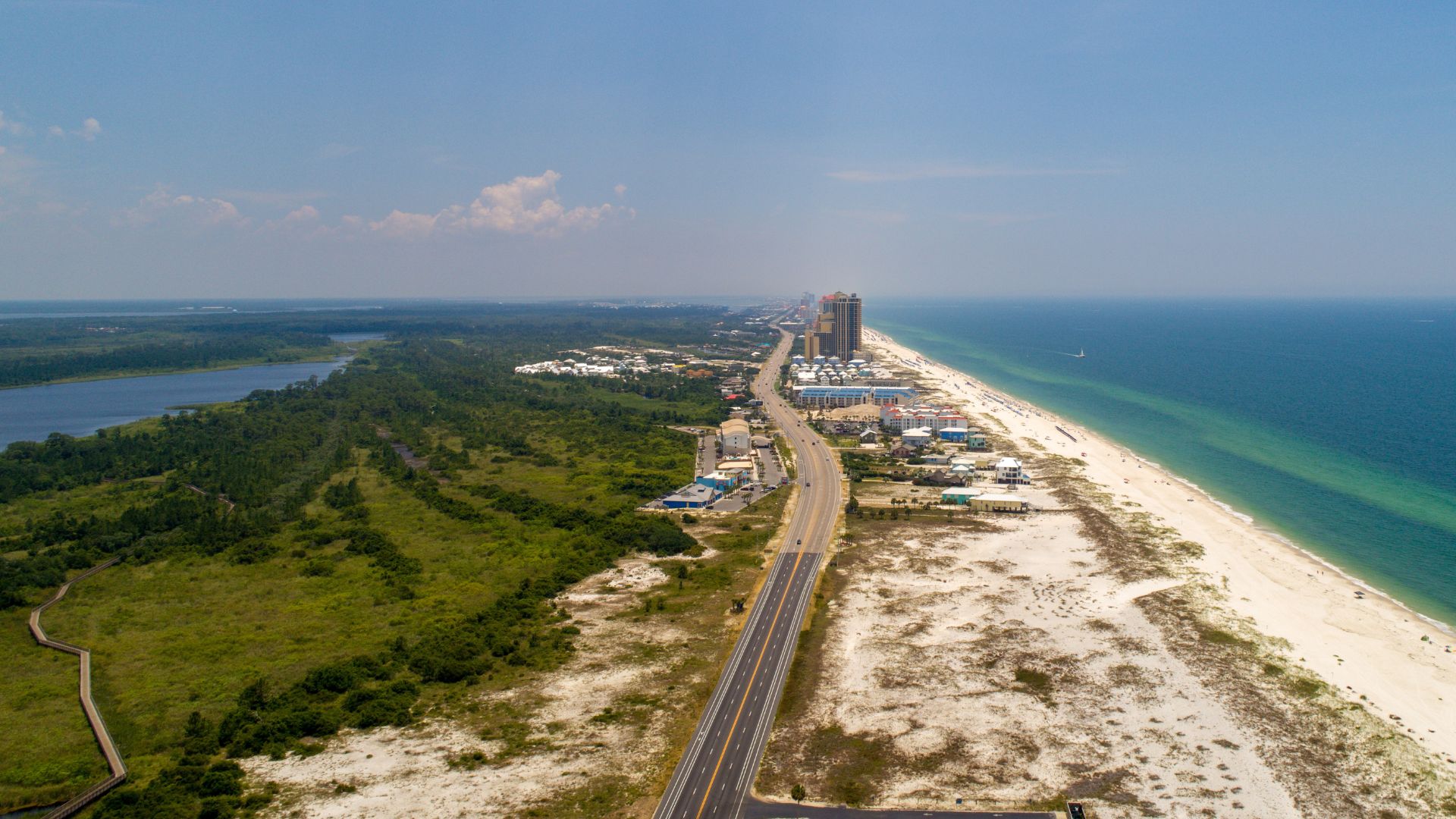 An aerial photograph of a coastal landscape featuring a long road and a strip of white sand beach separating the emerald water of the Gulf of Mexico from a lush green park area with a lake.